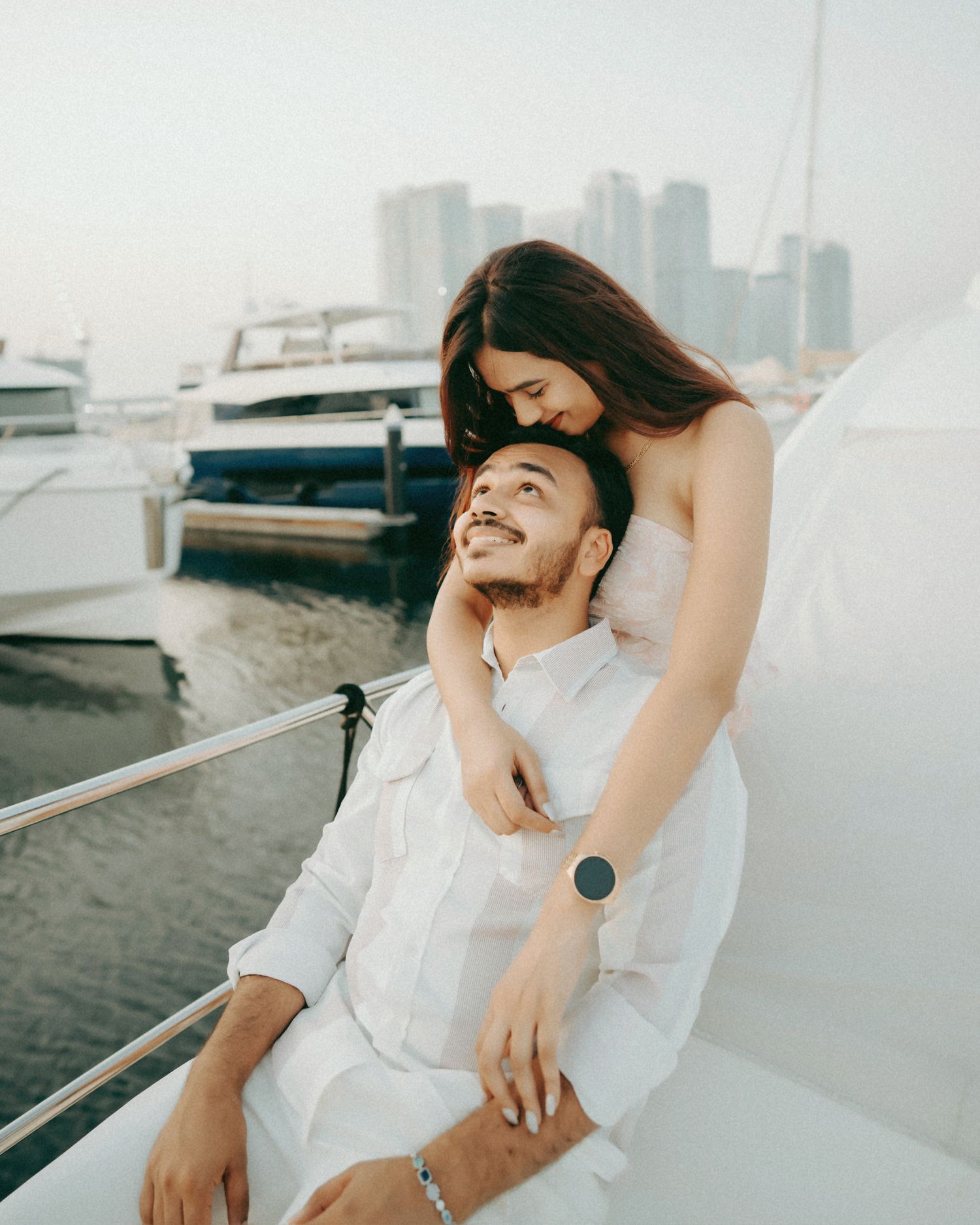 A couple in white outfits share a romantic moment on a yacht with a city skyline in the background.
