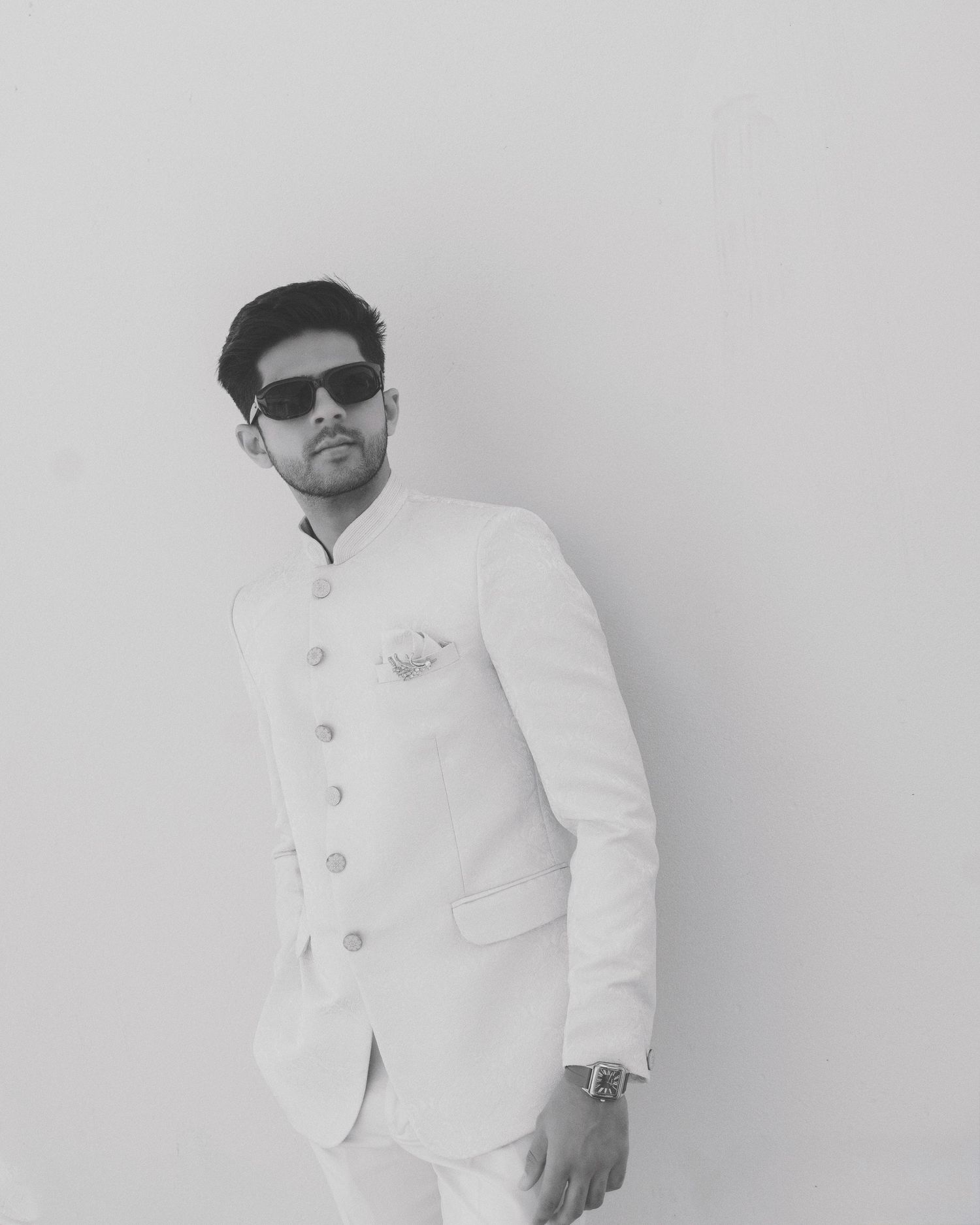Black and white photo of a stylish man in a white Nehru suit with sunglasses and a watch, posing confidently.