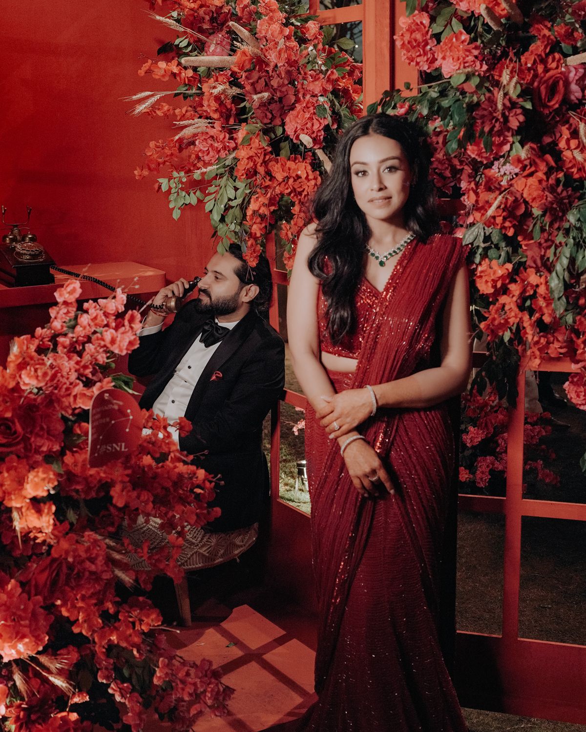 Elegant red saree portrait against vibrant floral backdrop with decorative lighting.