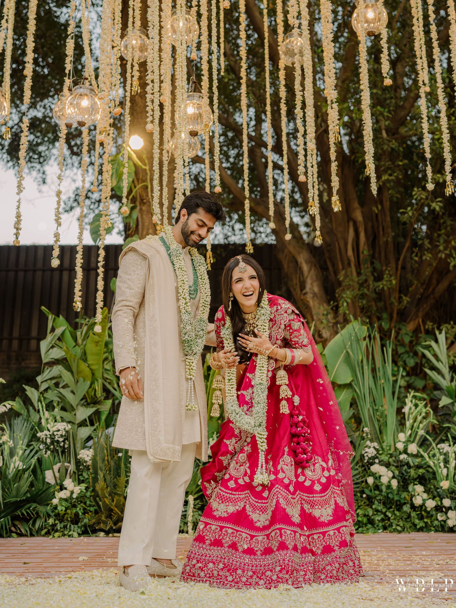 Indian couple in traditional wedding attire, groom in cream sherwani, bride in vibrant pink lehenga, under floral decor.