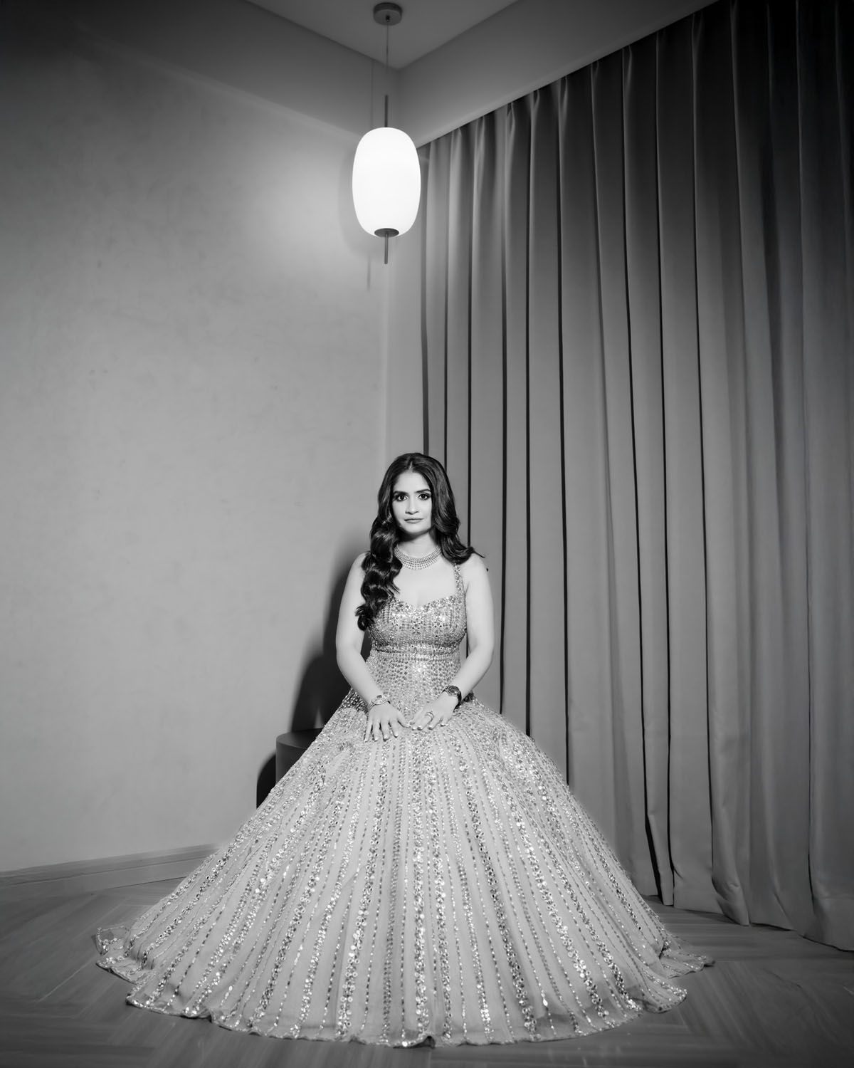 Elegant portrait of person in sparkly ball gown standing by curtains and pendant light.