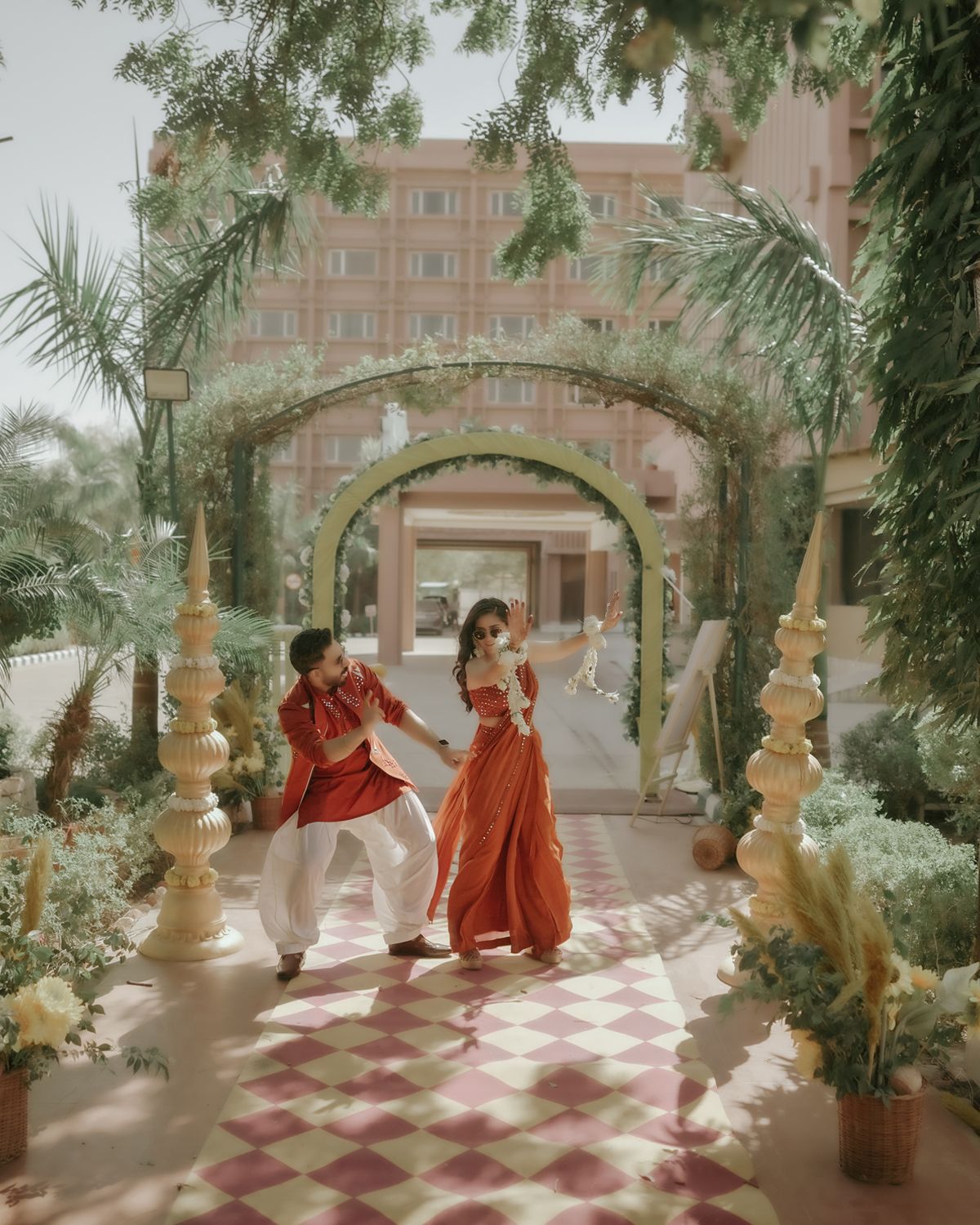 Couple in red traditional attire posing under a garden archway with decorative pillars.