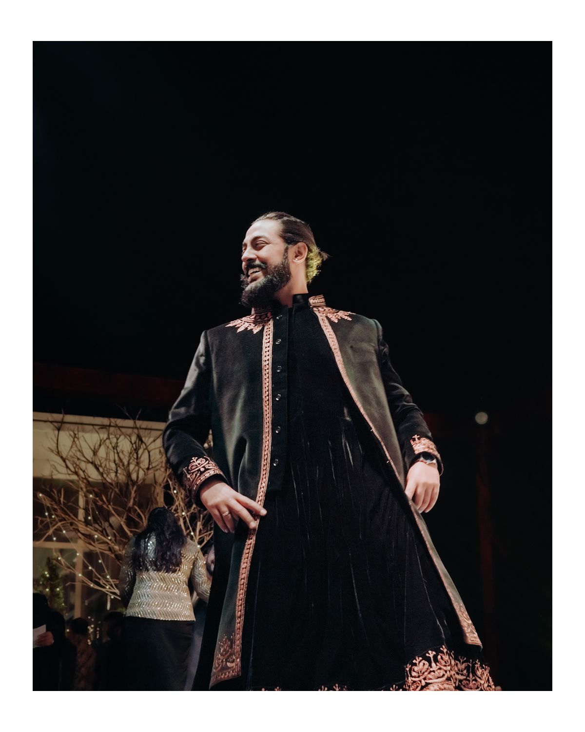 A performer in an ornate black velvet coat and embroidered details smiling during a theatrical production.