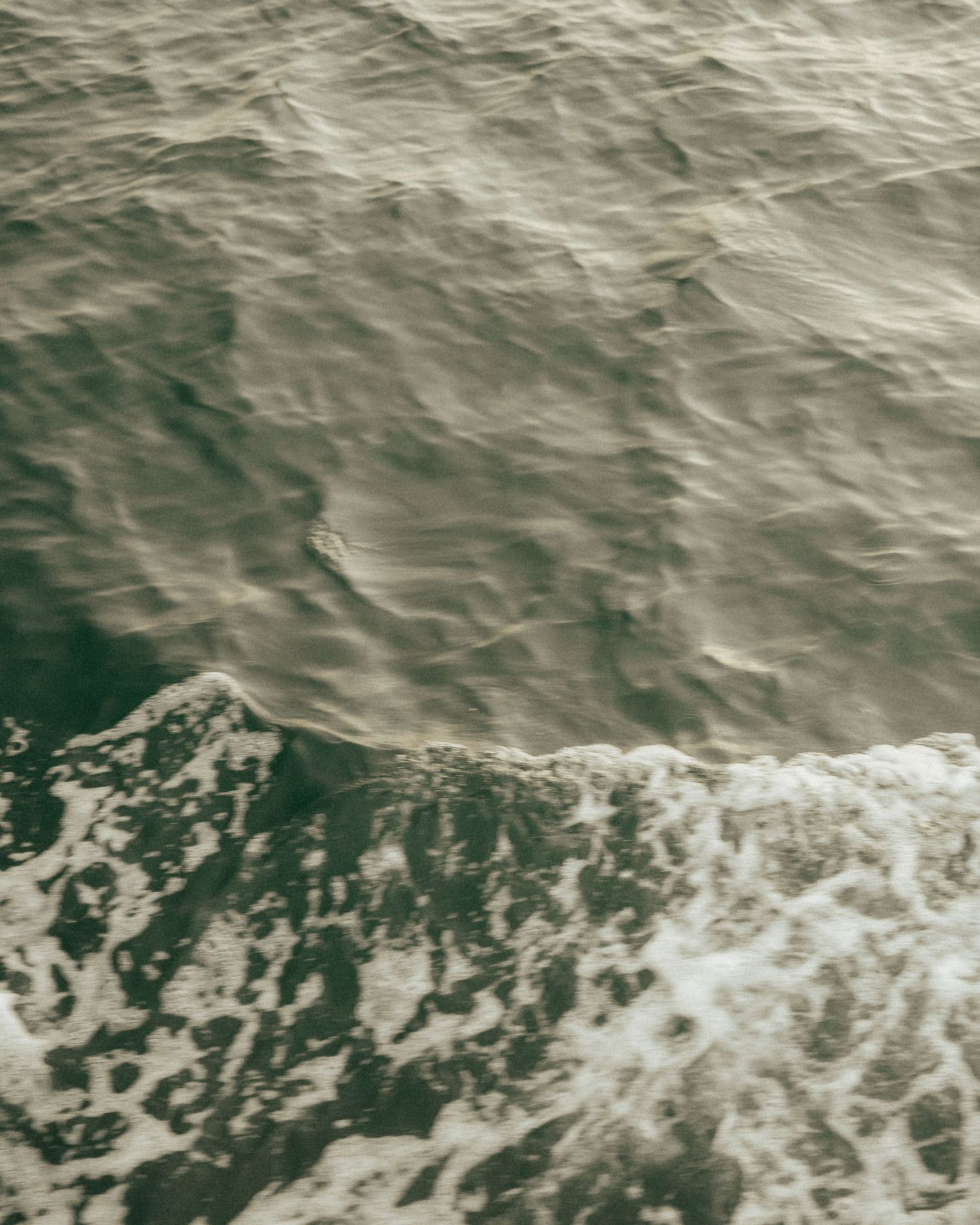 Aerial view of dark ocean waves with white sea foam crashing on the surface in muted green tones.