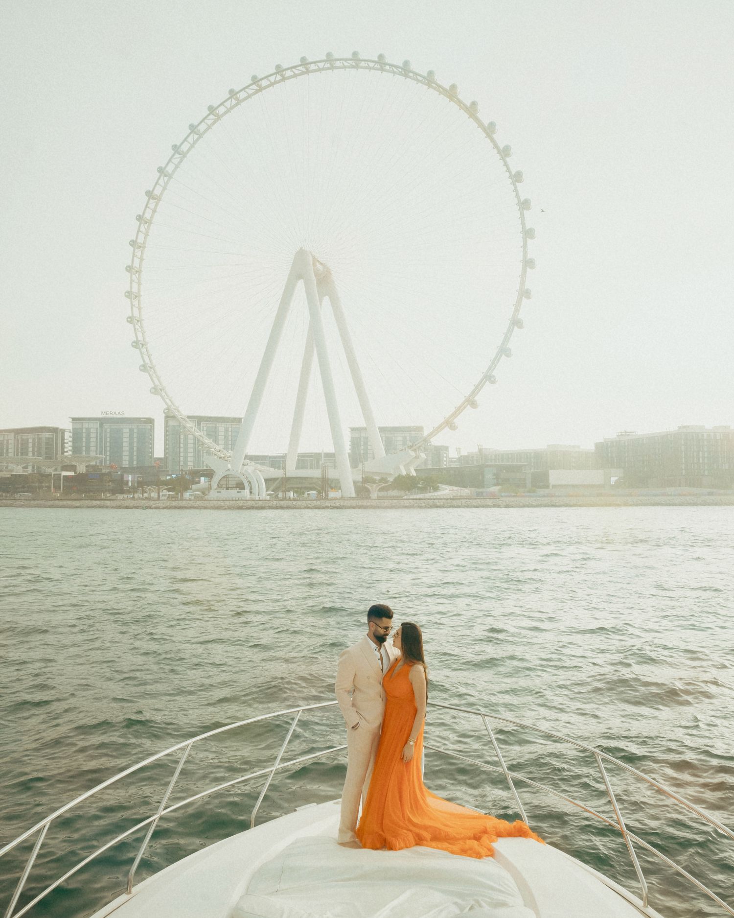 Couple in orange dress on yacht bow with Ain Dubai Ferris wheel backdrop at golden hour.