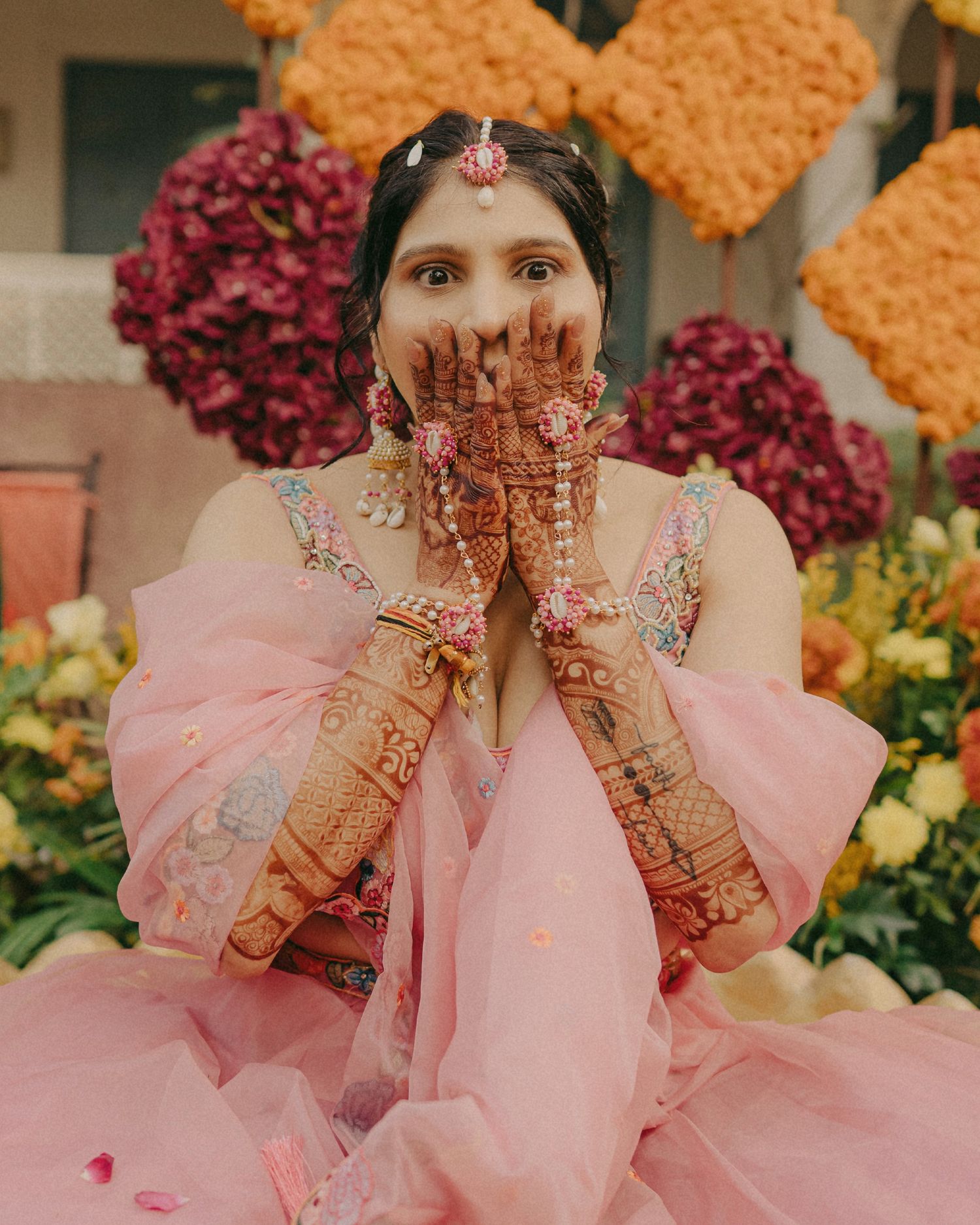 Bride in pink lehenga with intricate mehndi designs on hands, posing at floral backdrop during Indian wedding ceremony.