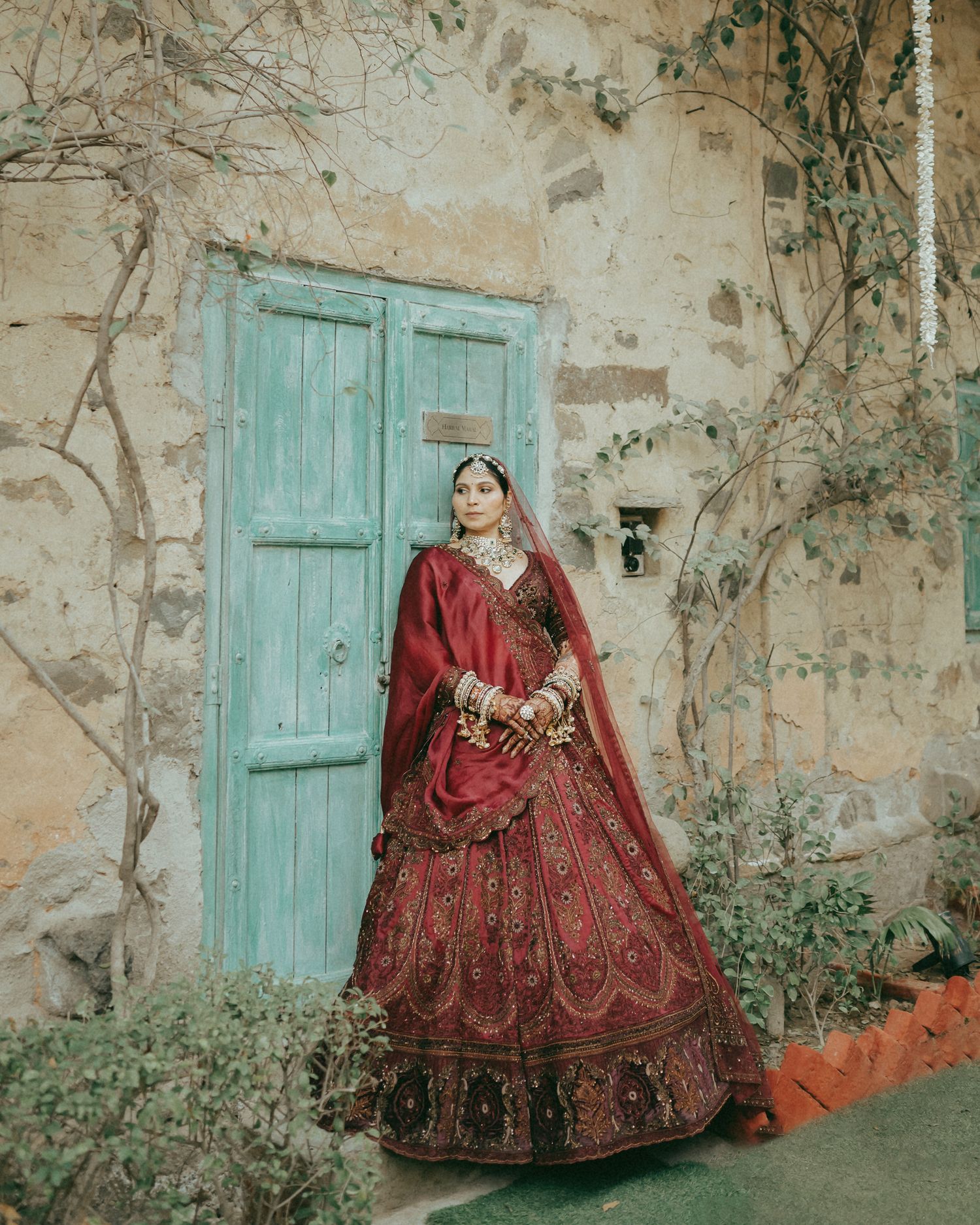 Bride in red bridal lehenga with gold embroidery poses before a turquoise door and rustic stone wall.
