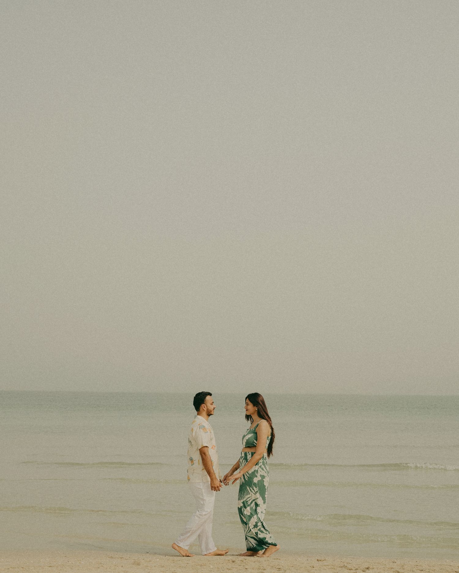Couple holding hands on a serene beach, man in white outfit, woman in green floral dress, ocean horizon backdrop.
