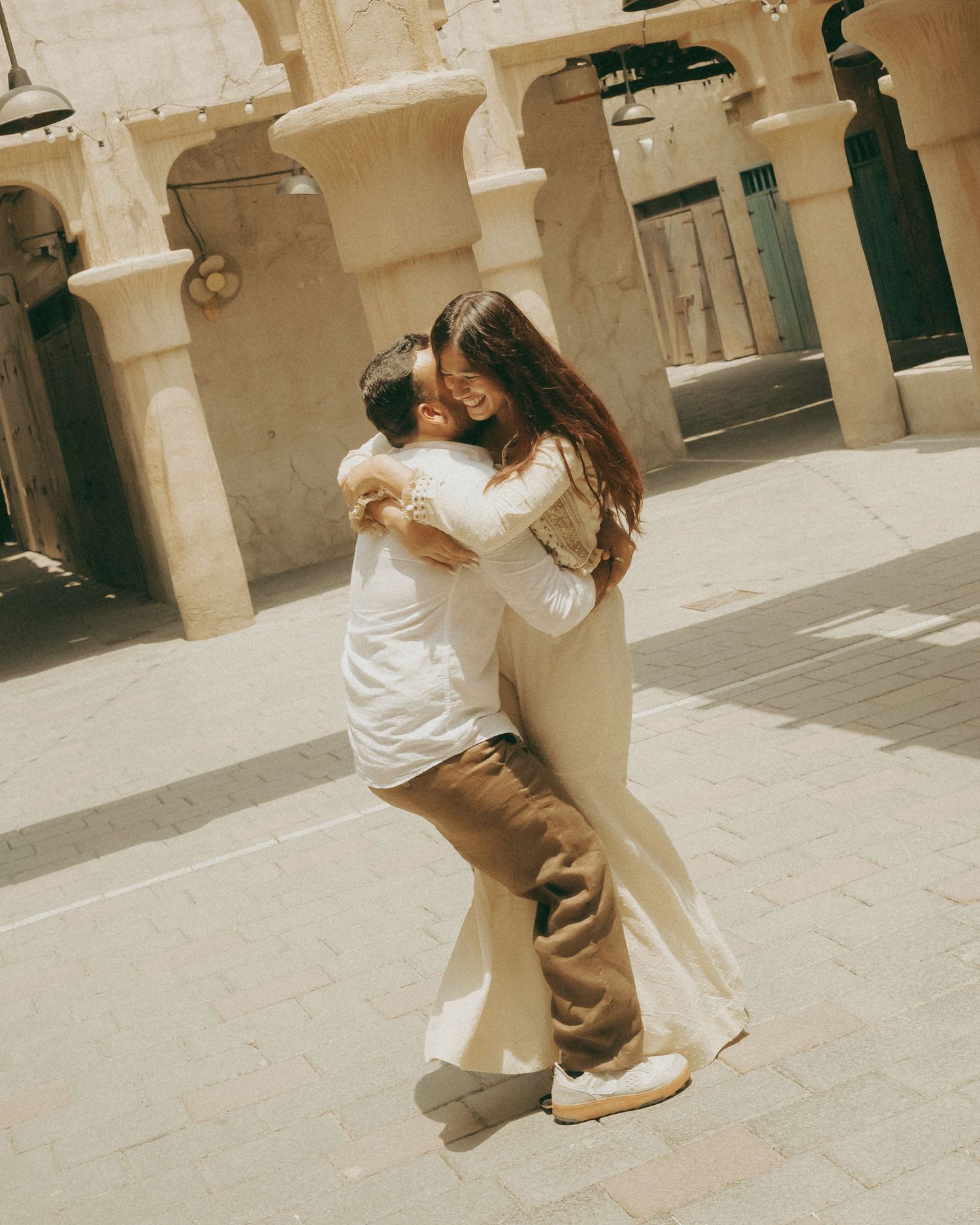 Couple embracing and kissing in a sunlit courtyard with beige stone archways in the background.