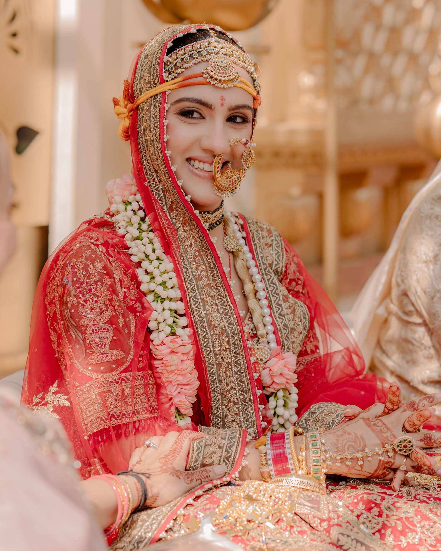 Smiling Indian bride in red and gold bridal lehenga with floral jewelry and traditional nose ring.