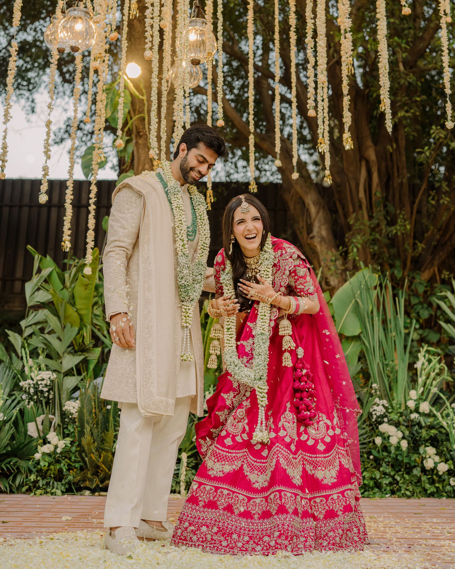 Couple in traditional Indian wedding attire, bride in pink lehenga, groom in beige sherwani, posing under floral decor.