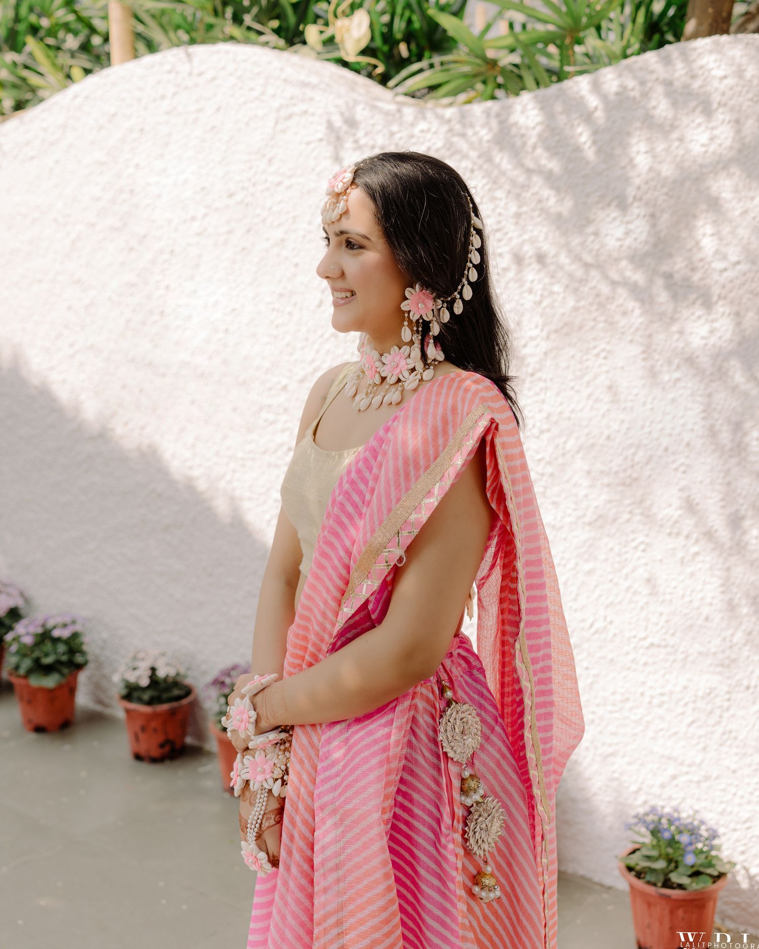 A woman in a pink striped lehenga with floral jewelry poses gracefully outdoors near potted plants.