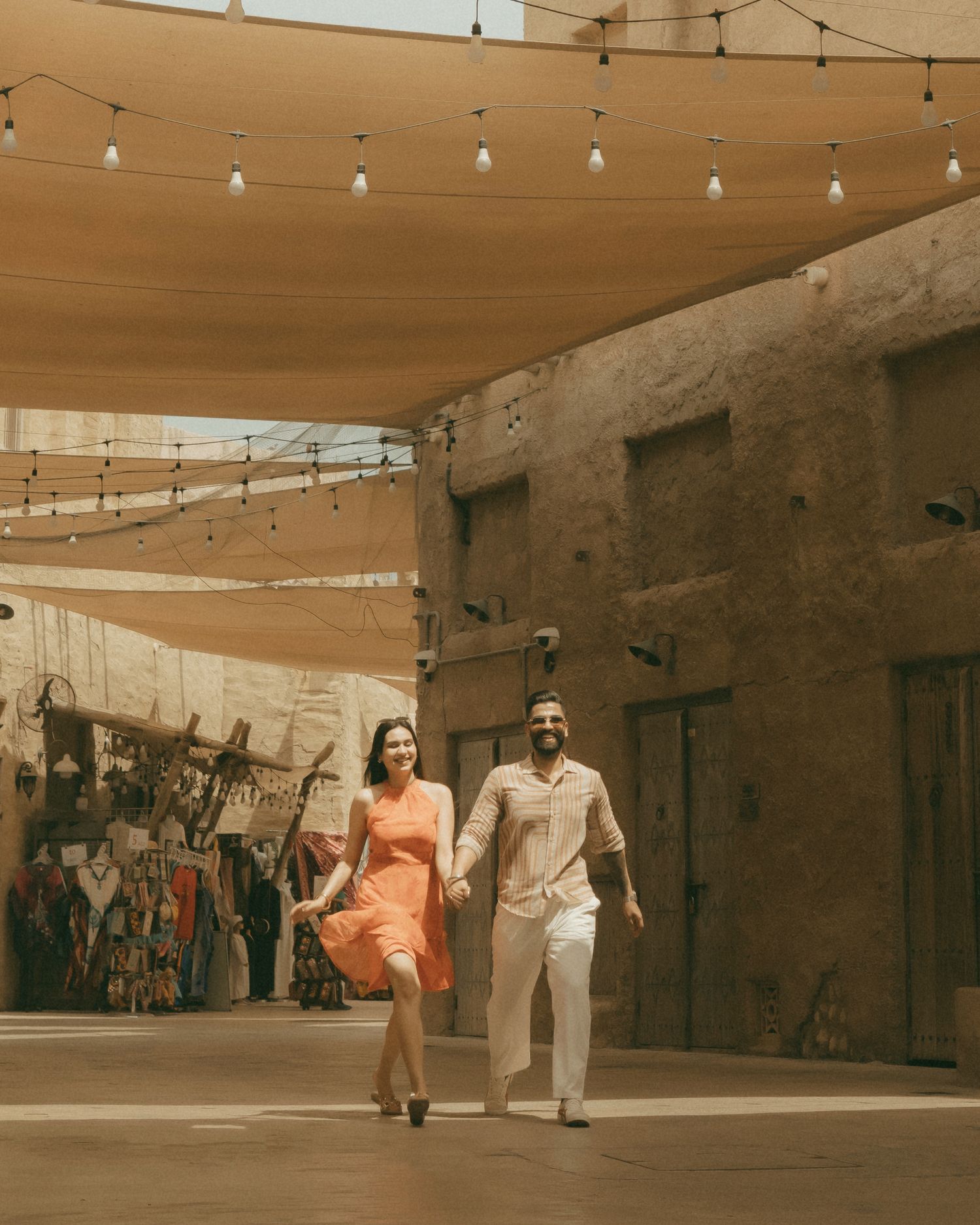 Couple holding hands walking under string lights and shade sails in a warm-toned outdoor market setting.