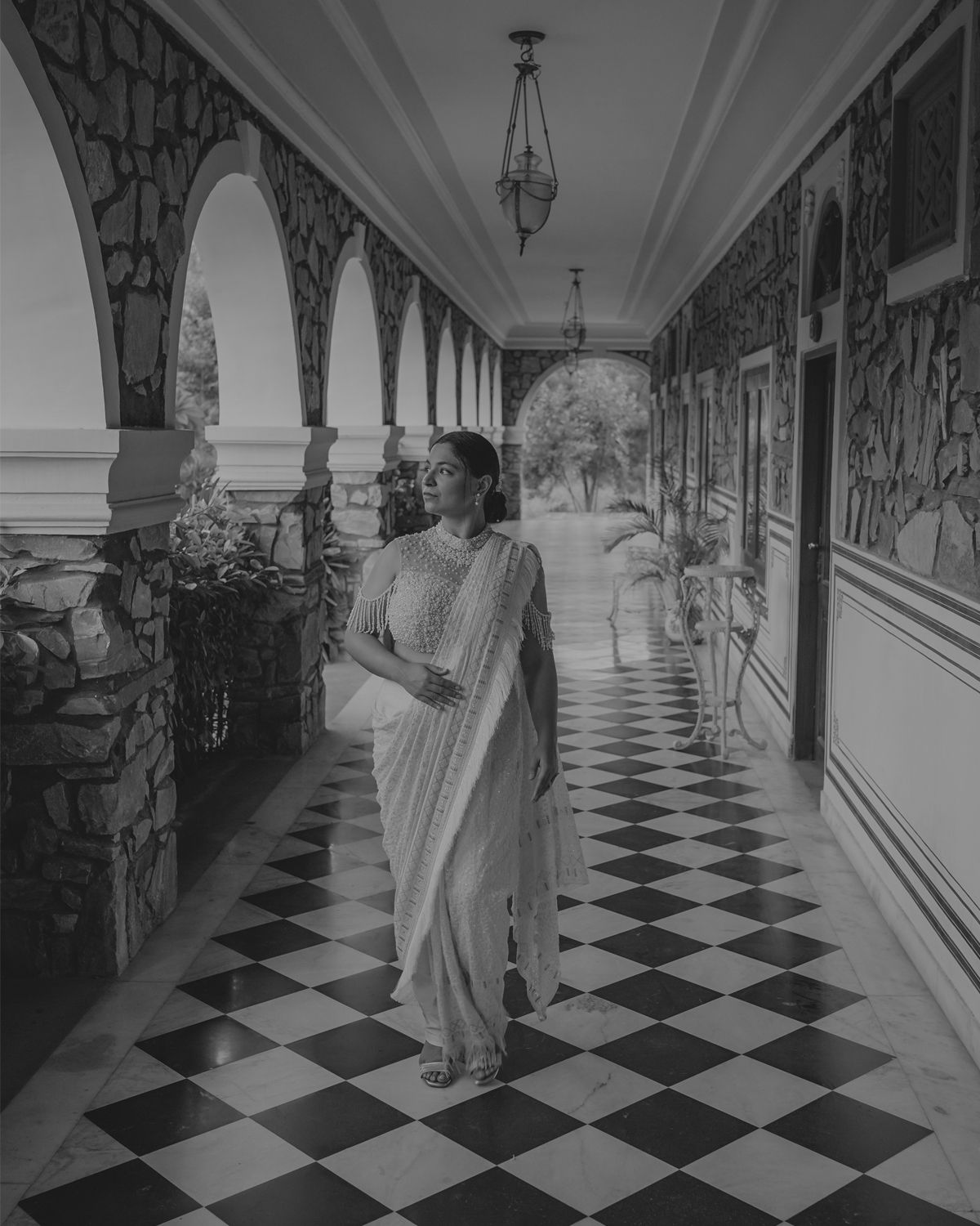 A traditional Indian saree-clad figure poses elegantly in a hallway with striking black and white checkered floors and arched columns.