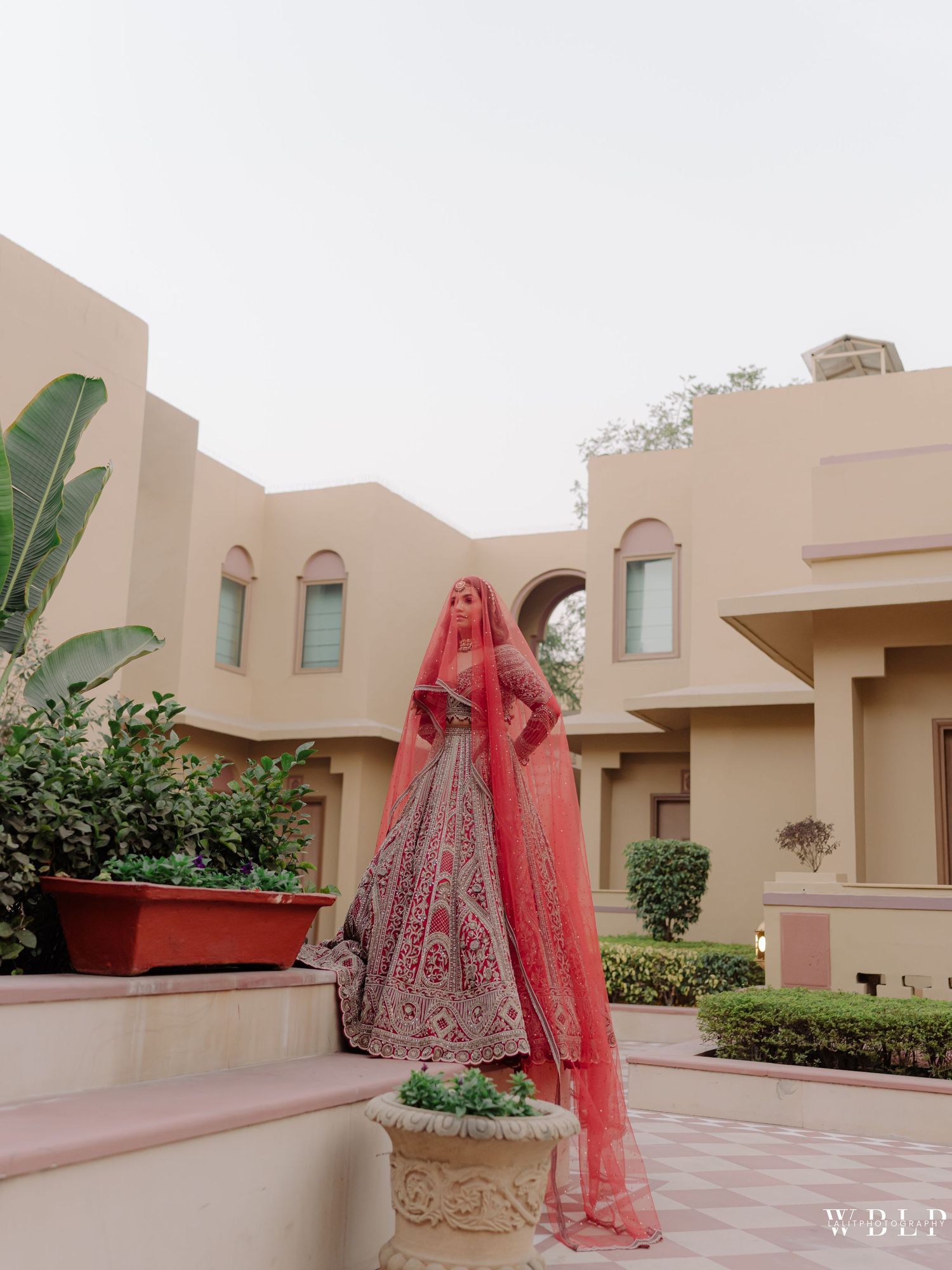 Bride in red bridal lehenga with dupatta poses at elegant beige heritage venue outdoors.
