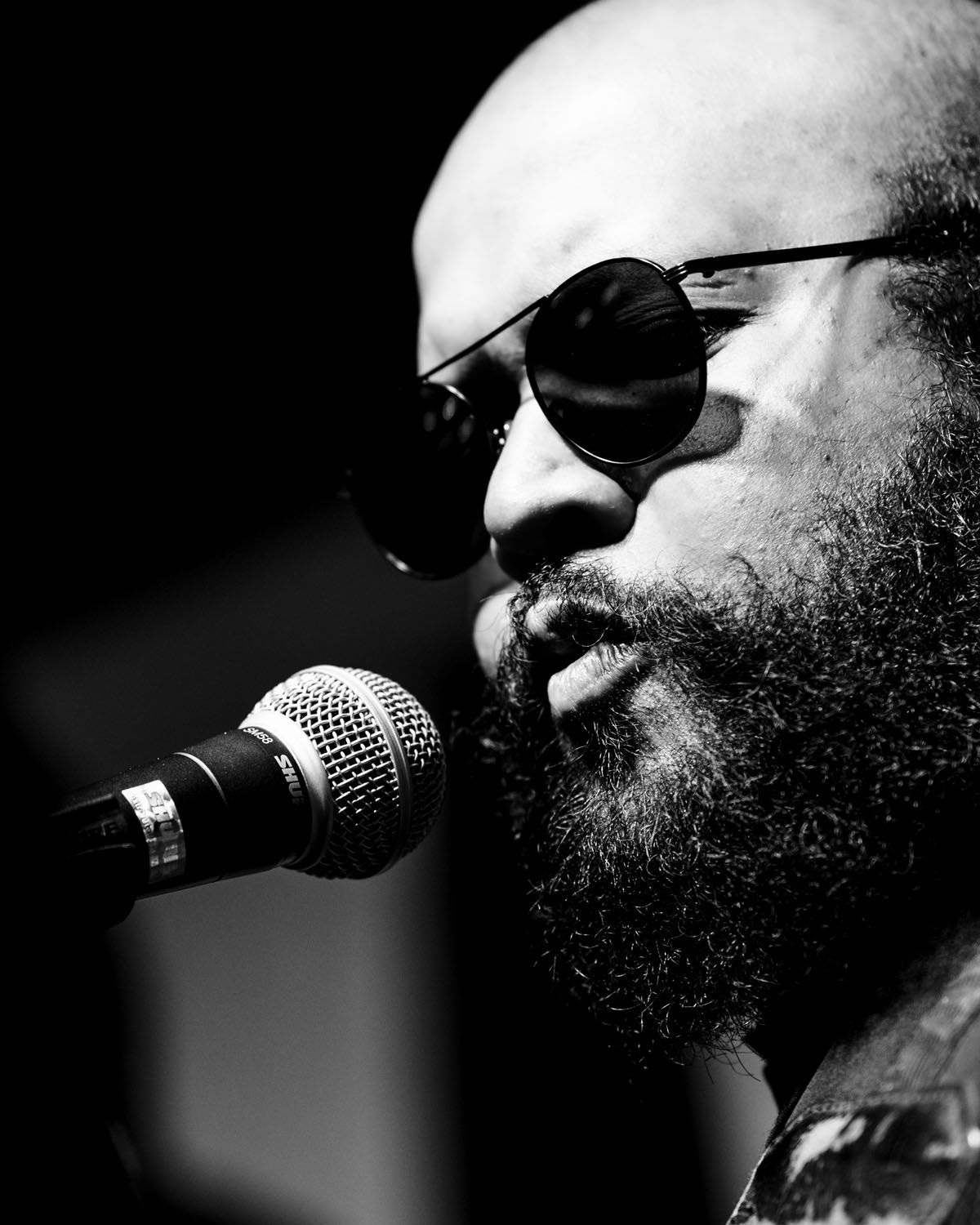 Black and white profile shot of a musician with a full beard and sunglasses singing into a microphone.
