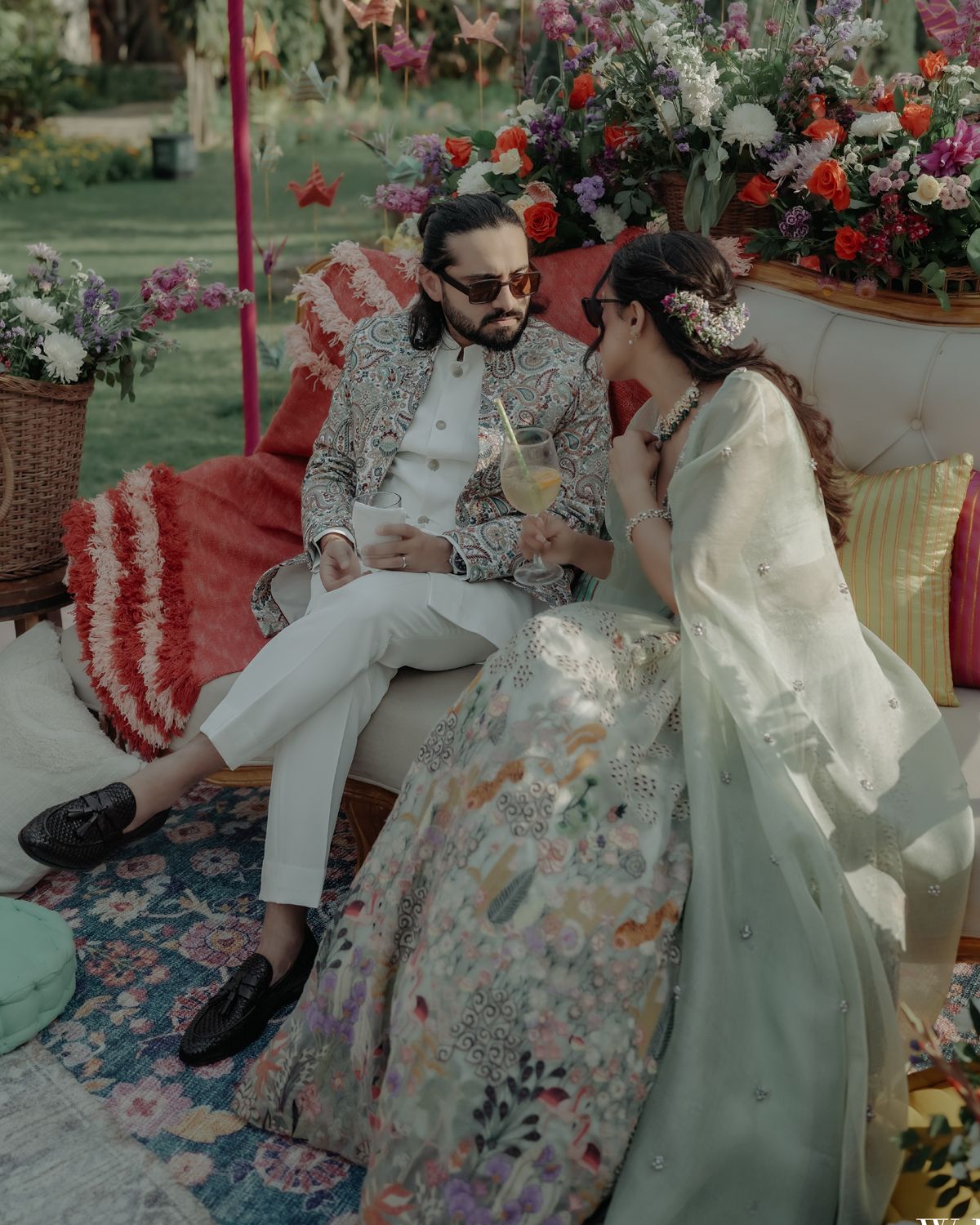 Elegantly dressed couple sits together on floral cushions at outdoor garden party surrounded by red and white flowers.