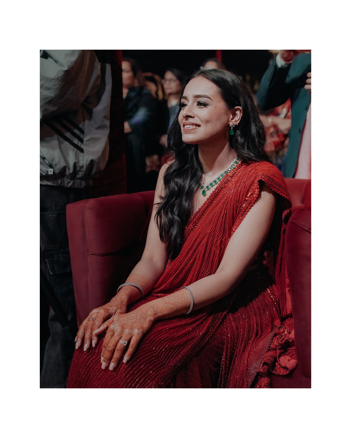 Person in an elegant red pleated dress sits comfortably on a burgundy armchair at a formal event.
