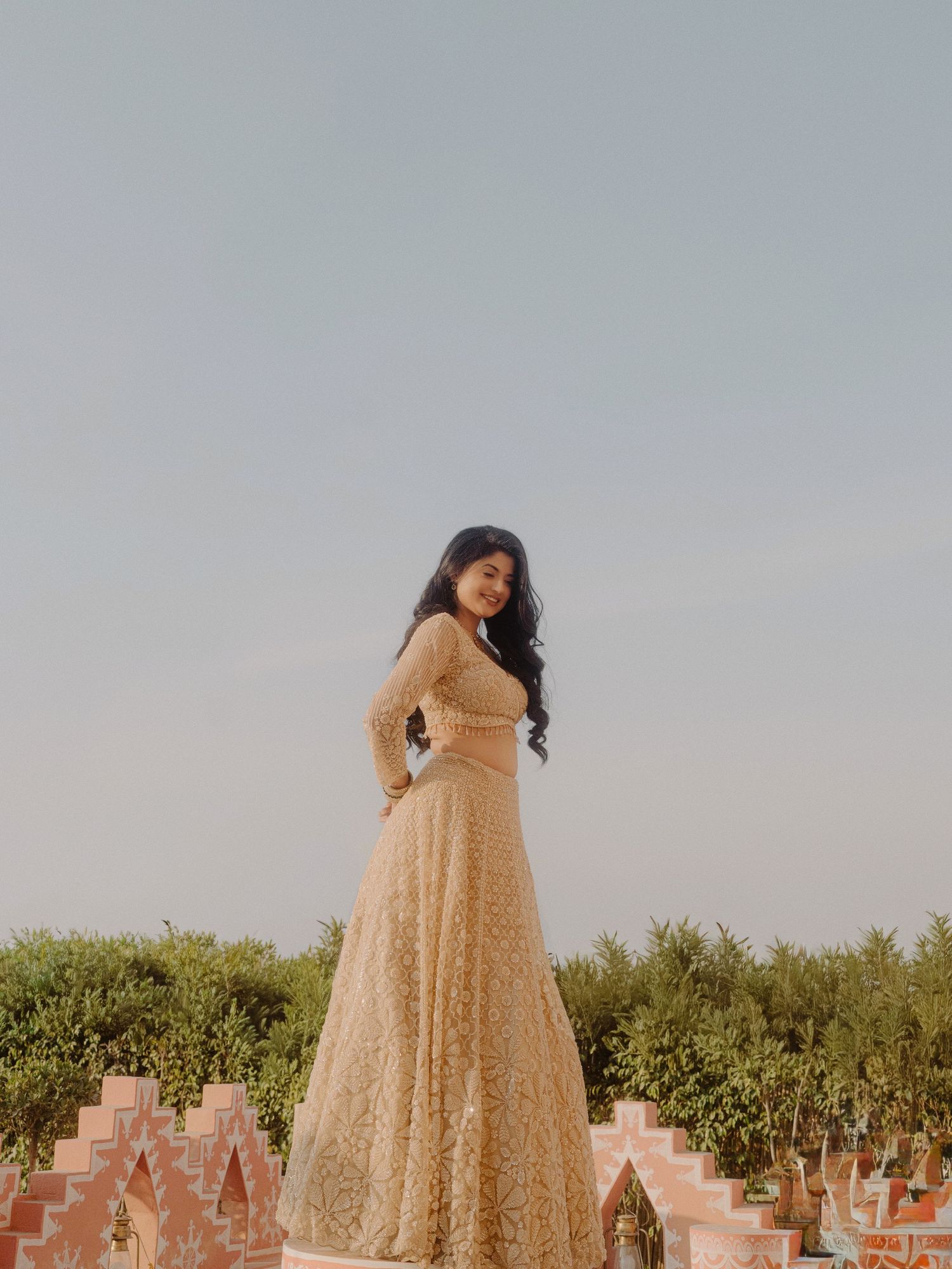 A young woman in an elegant golden lace lehenga poses gracefully outdoors against a clear sky with lush greenery.