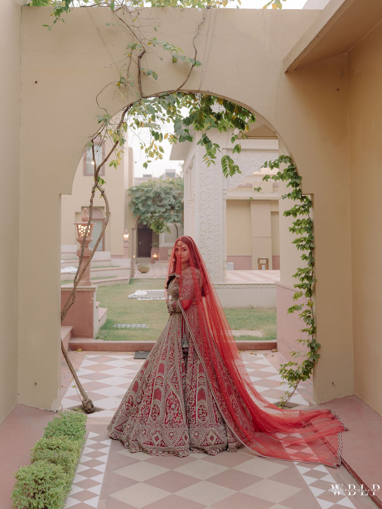 Bride in red and grey embroidered lehenga with dupatta stands under arched doorway at elegant Indian wedding venue.