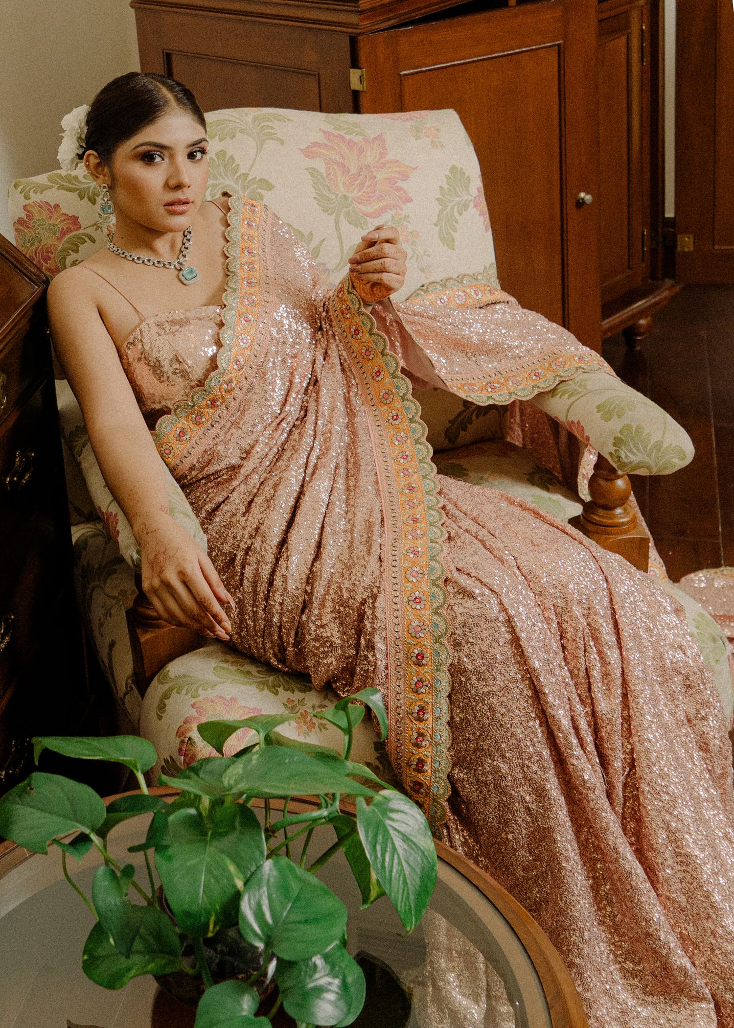Elegant South Asian formal wear in rose gold sequins styled with indoor plants and antique wooden furniture in vintage setting.