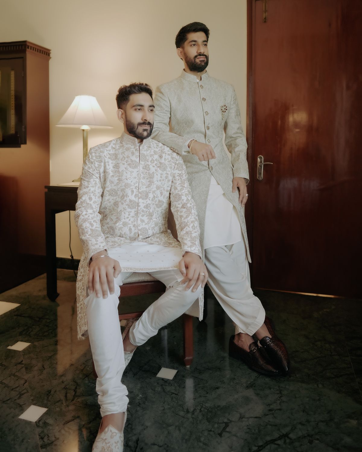Two men wearing matching light-colored Indian formal attire pose in an indoor setting.