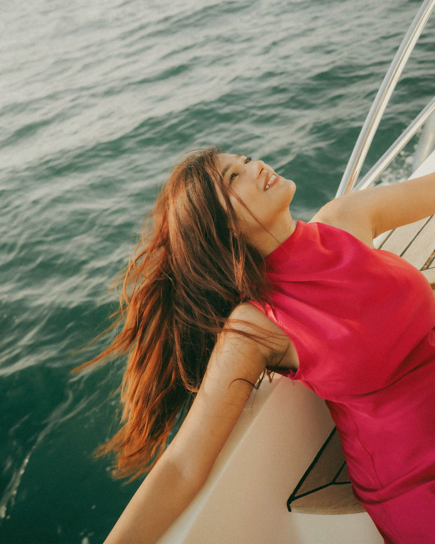 Woman in pink dress leaning back on sailboat bow, hair blowing in wind over turquoise ocean waters.