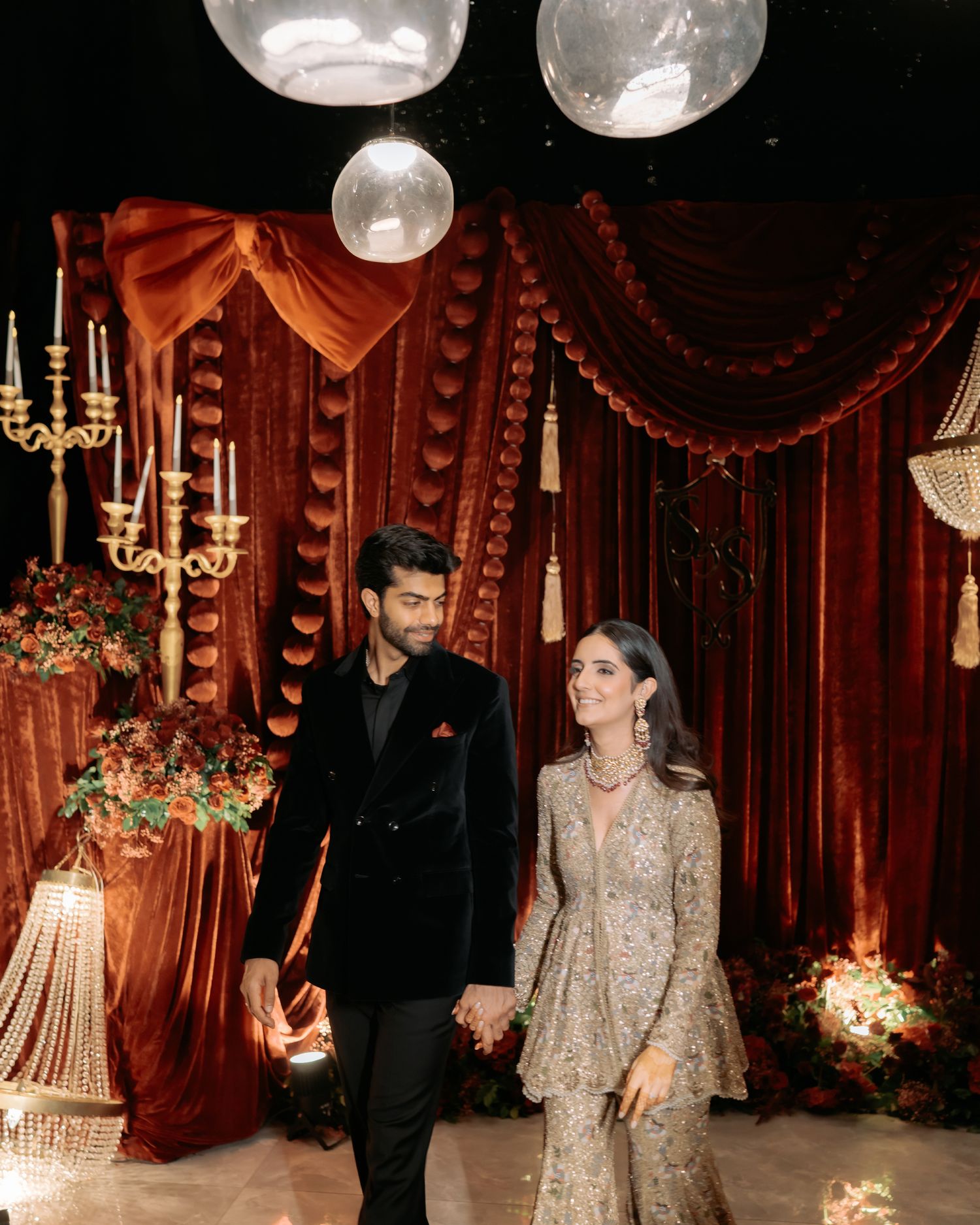 Elegant couple in formal attire holding hands at a luxurious wedding venue with red velvet drapes and candlelight.
