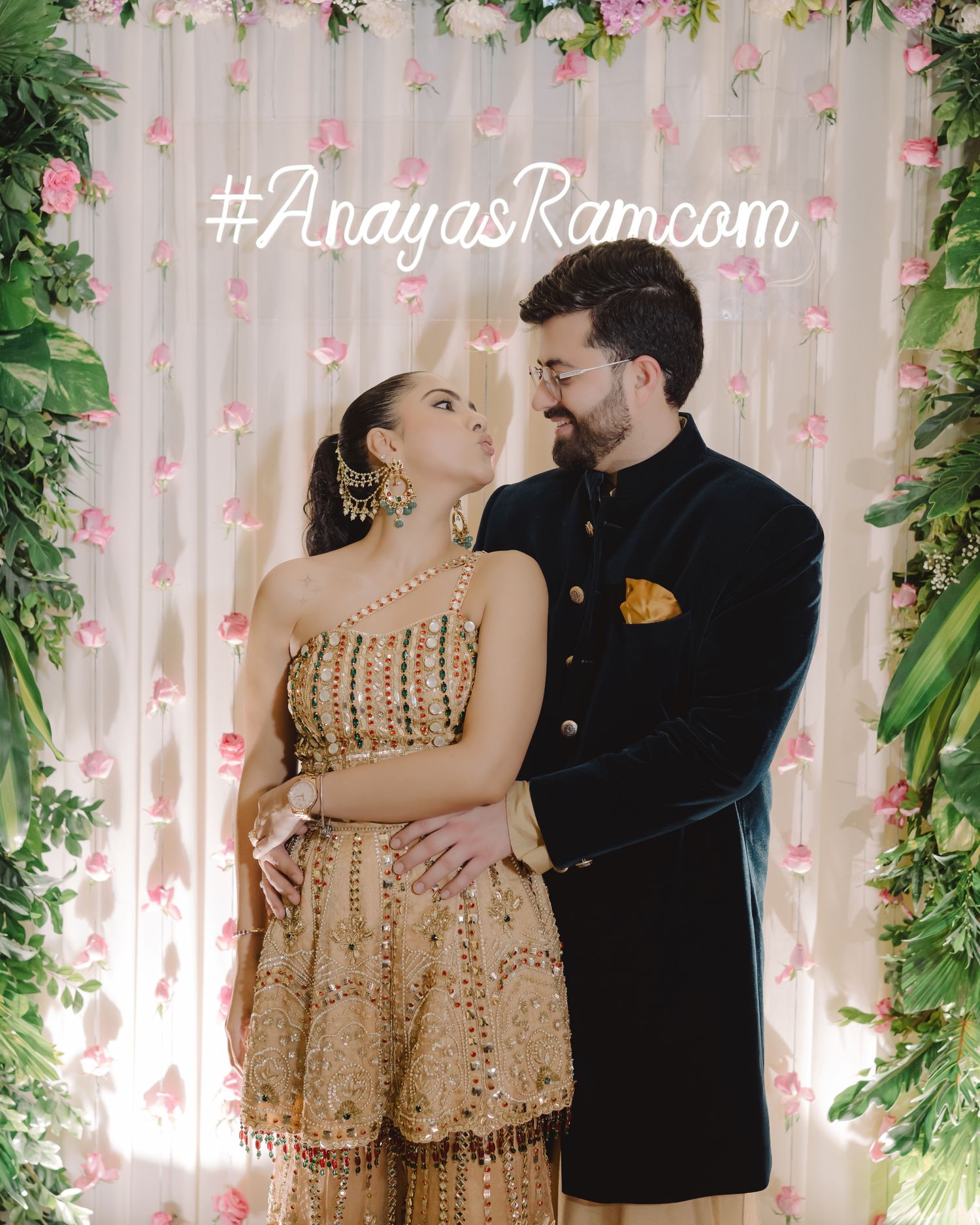 Couple in Indian wedding attire posing before floral backdrop with #AnayasRamcom hashtag.