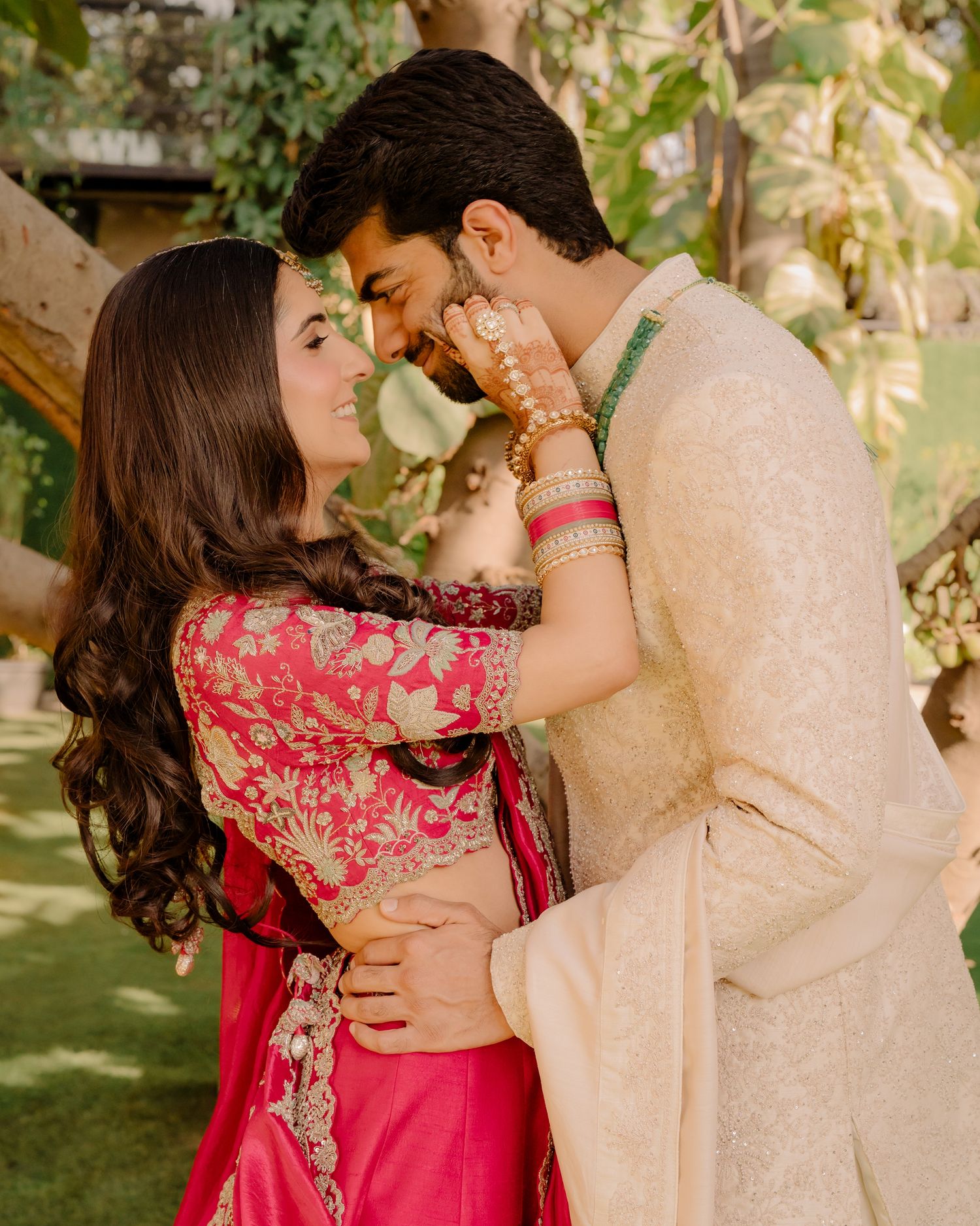 Couple in traditional Indian attire sharing a romantic moment outdoors, woman in pink lehenga, man in cream sherwani.