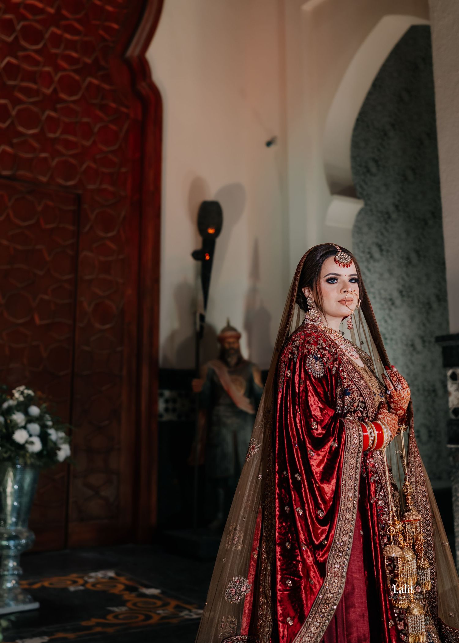 Bride wearing deep red velvet lehenga poses in an ornate architectural setting with arched doorways.