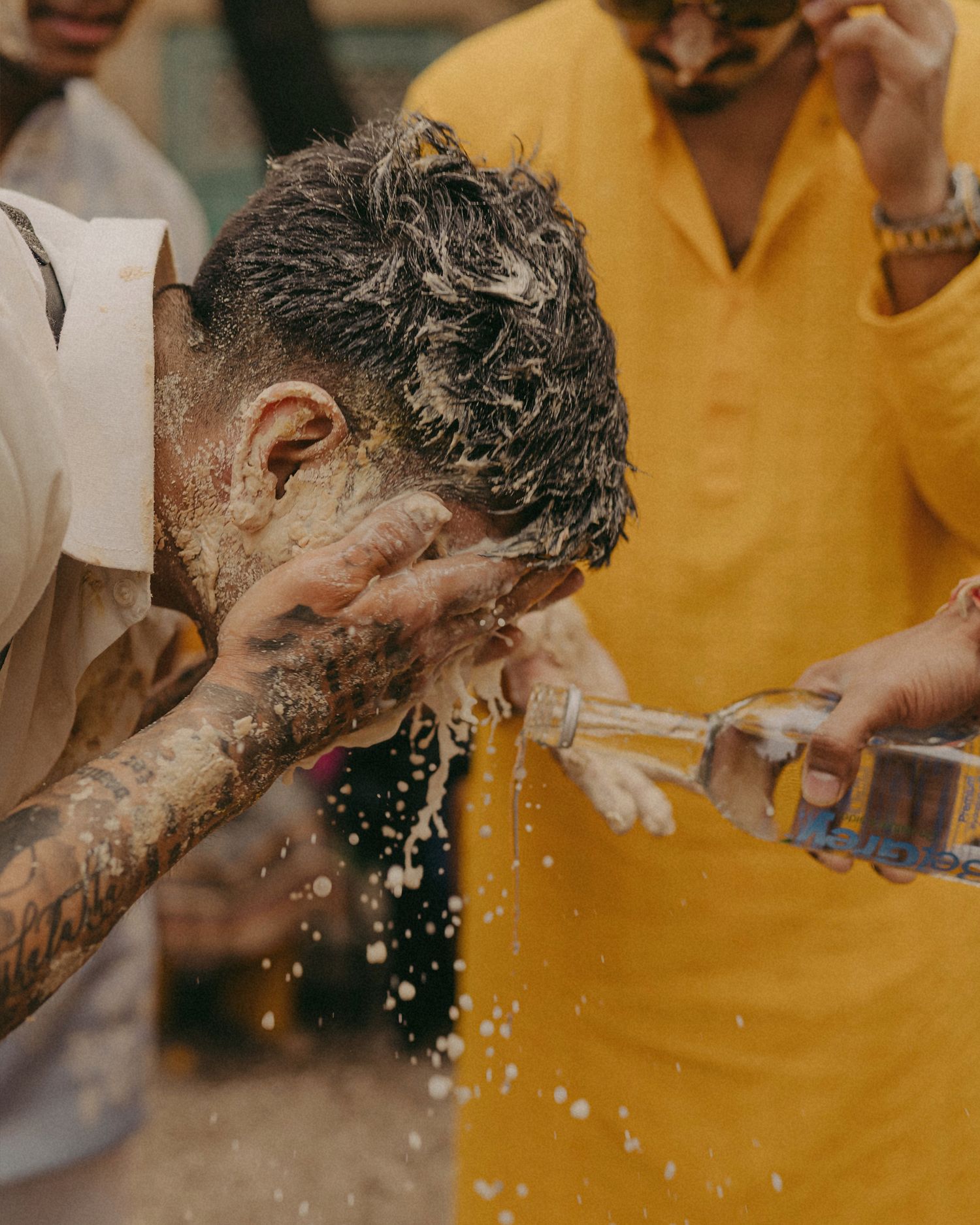 Tattooed man washing his face with water poured from a bottle outdoors, with a person in yellow clothing nearby.