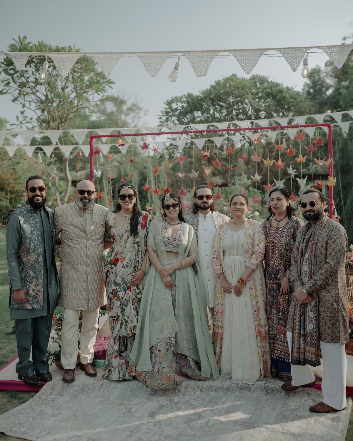 A group in traditional Indian wedding attire poses together in front of a decorated backdrop with white bunting overhead.