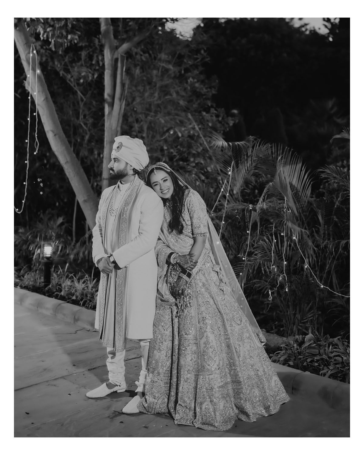 A couple stands together in traditional South Asian wedding attire including a lengha and sherwani in a garden setting.