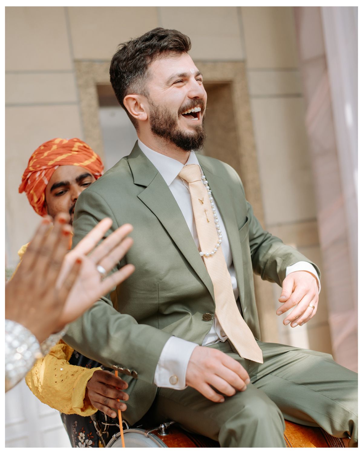 Wedding guests share a joyful moment at an outdoor celebration, with one wearing a sage green suit and orange tie.