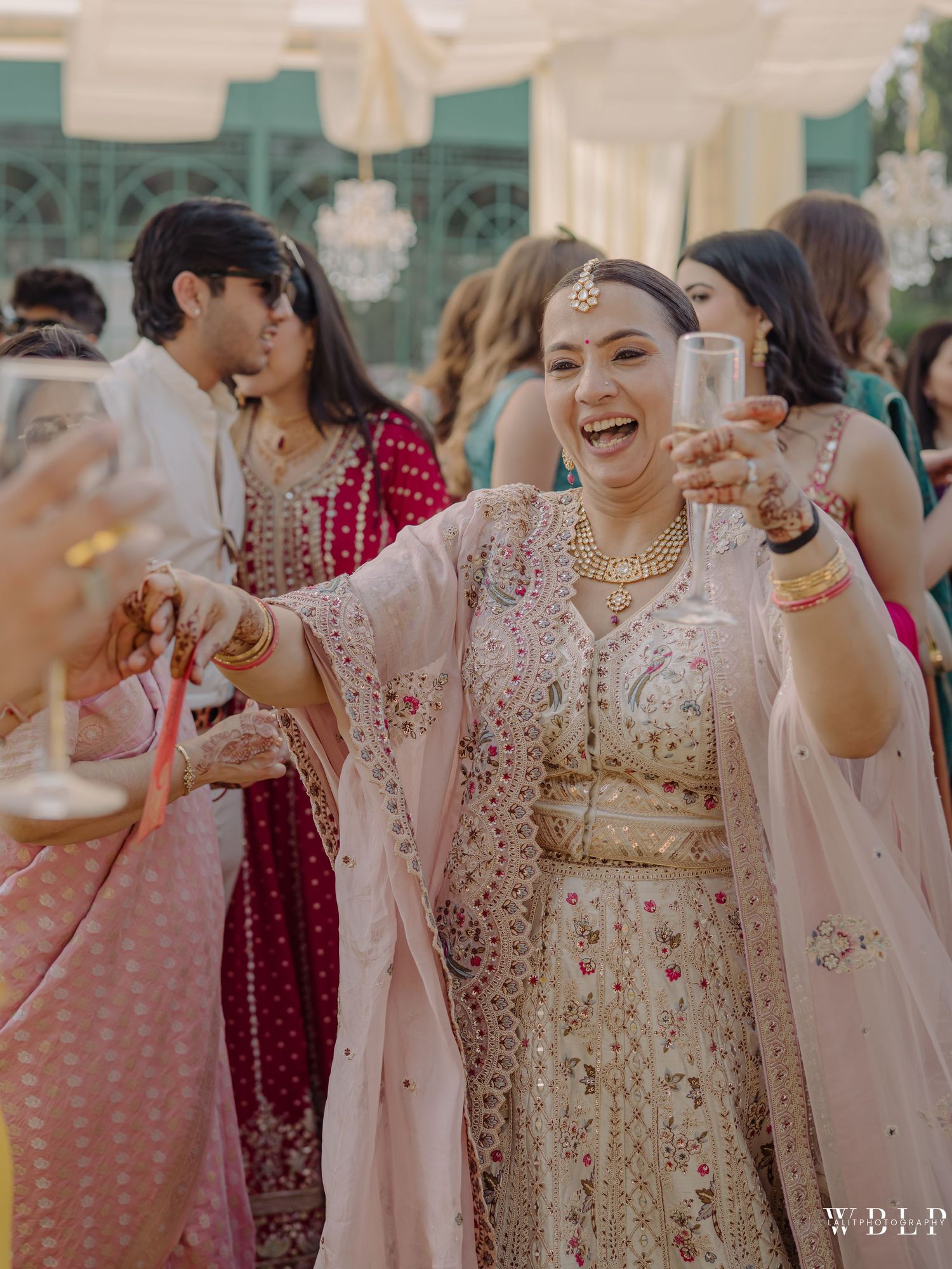 Joyful bride in gold lehenga and pink dupatta toasting champagne at an Indian wedding celebration.