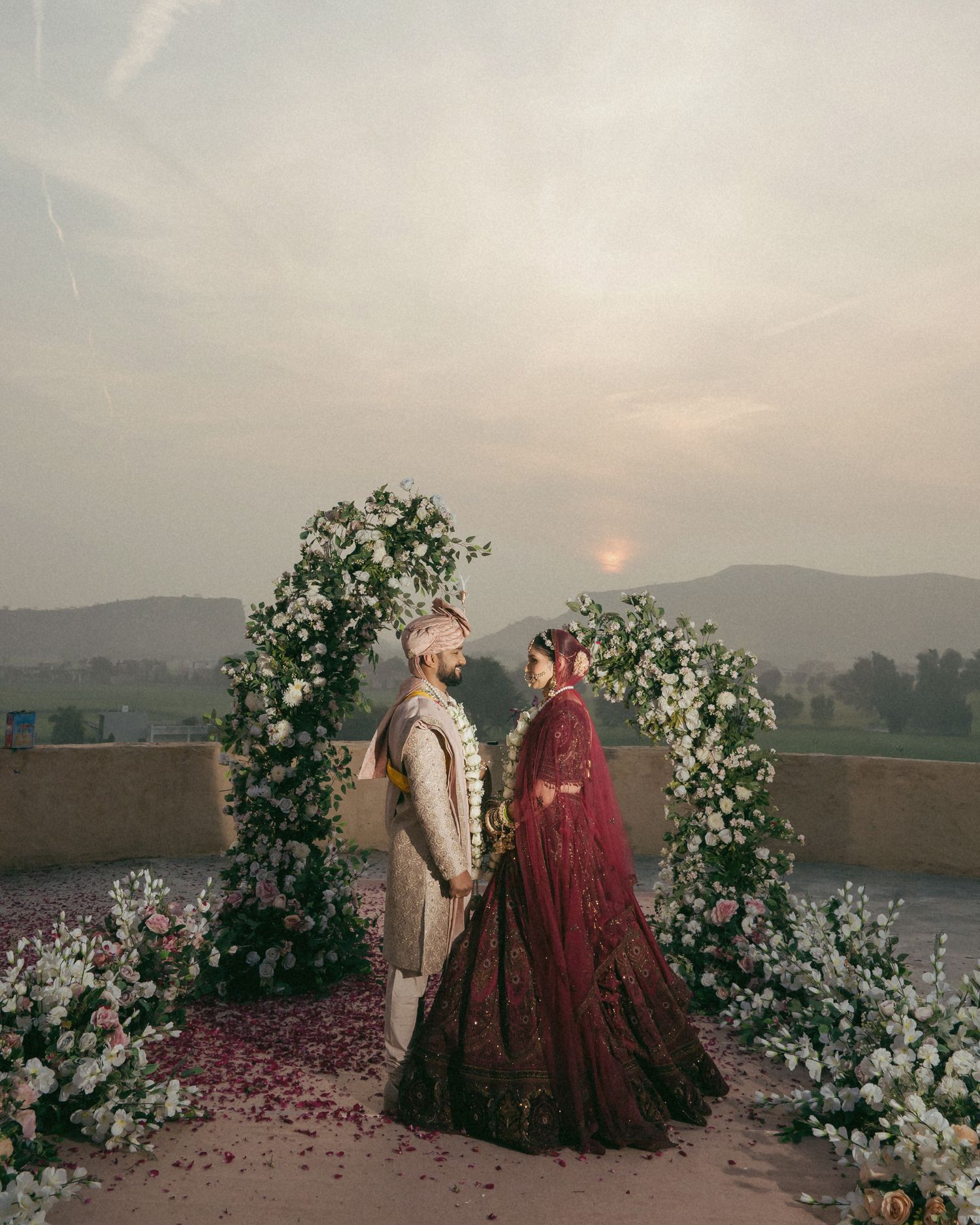 Indian couple in traditional wedding attire standing under floral arch at sunset with mountain backdrop.
