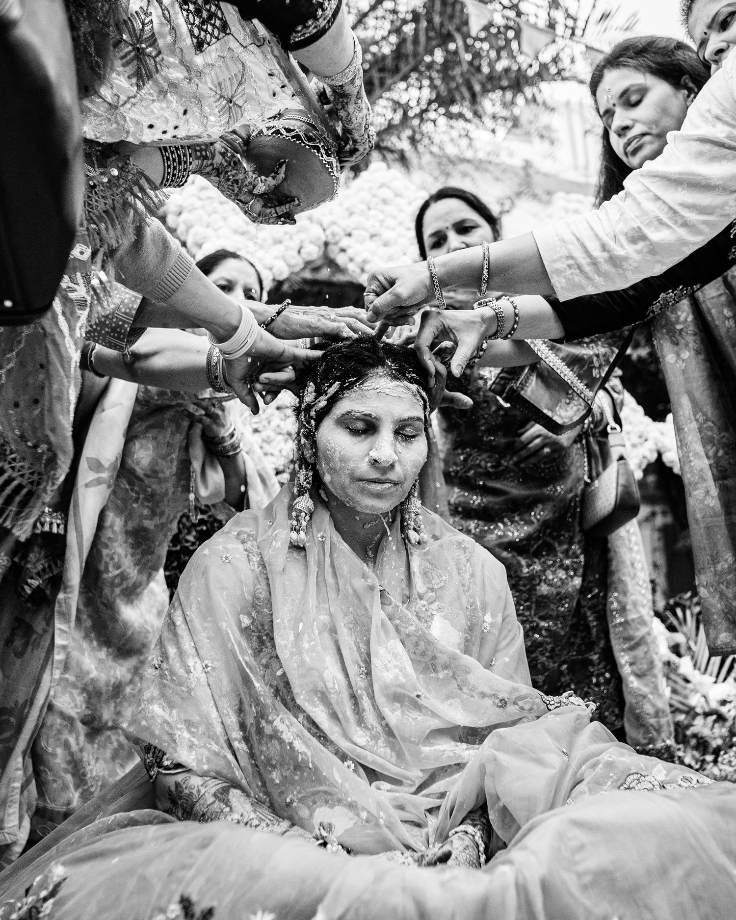 Black and white photo of a bride seated during a traditional South Asian wedding ceremony with family members surrounding her.