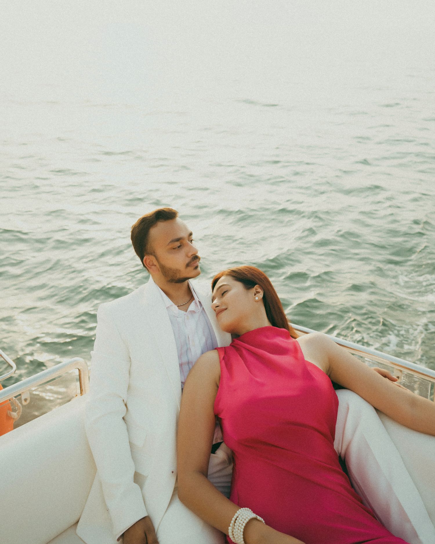 A couple in elegant attire relaxing on a boat with ocean backdrop, man in white suit and woman in pink dress.