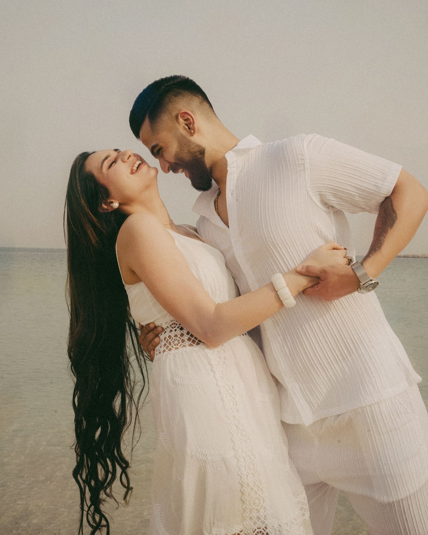 Couple in matching white outfits embracing and laughing on a beach with calm ocean water in the background.