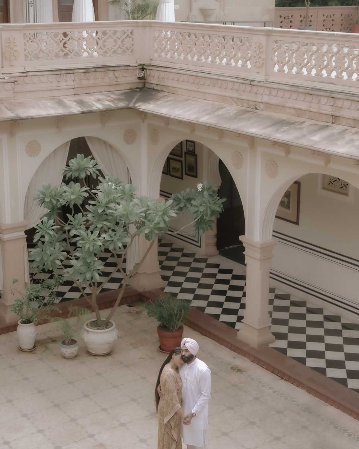 Two figures stand in an elegant courtyard with potted plants and a checkerboard floor pattern beneath ornate arches.