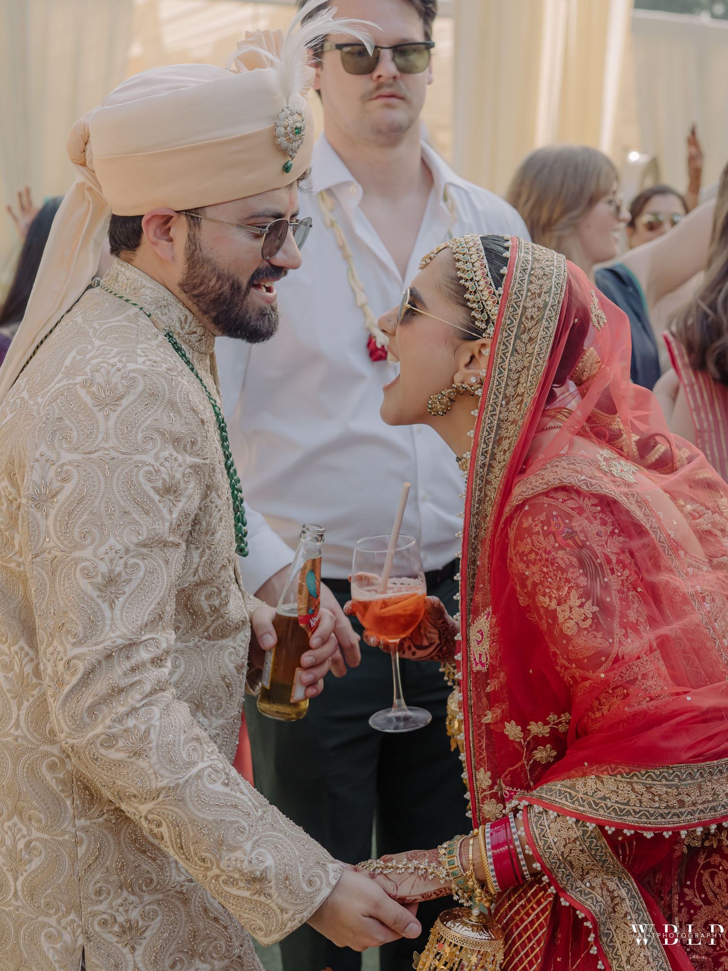 Indian bride in red lehenga and groom in ivory sherwani share a joyful moment with drinks at their wedding celebration.