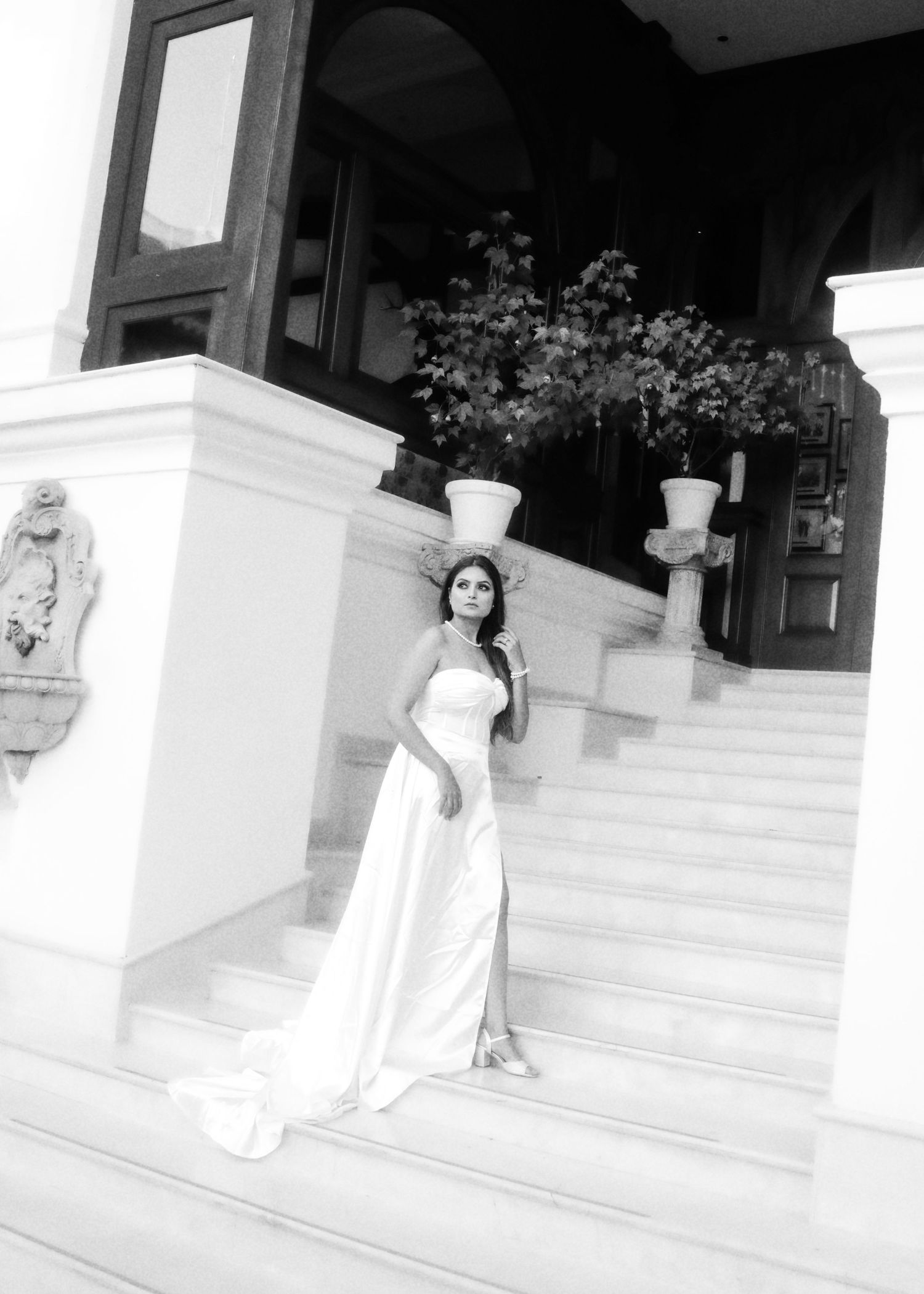 A bride in a flowing white gown poses elegantly on marble steps outside an ornate building entrance in black and white.