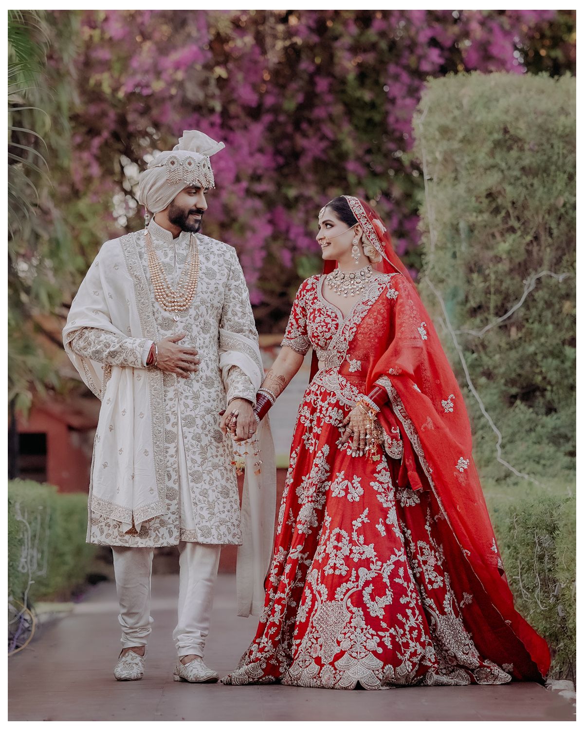 Couple in traditional Indian wedding attire posing against a backdrop of pink flowers.