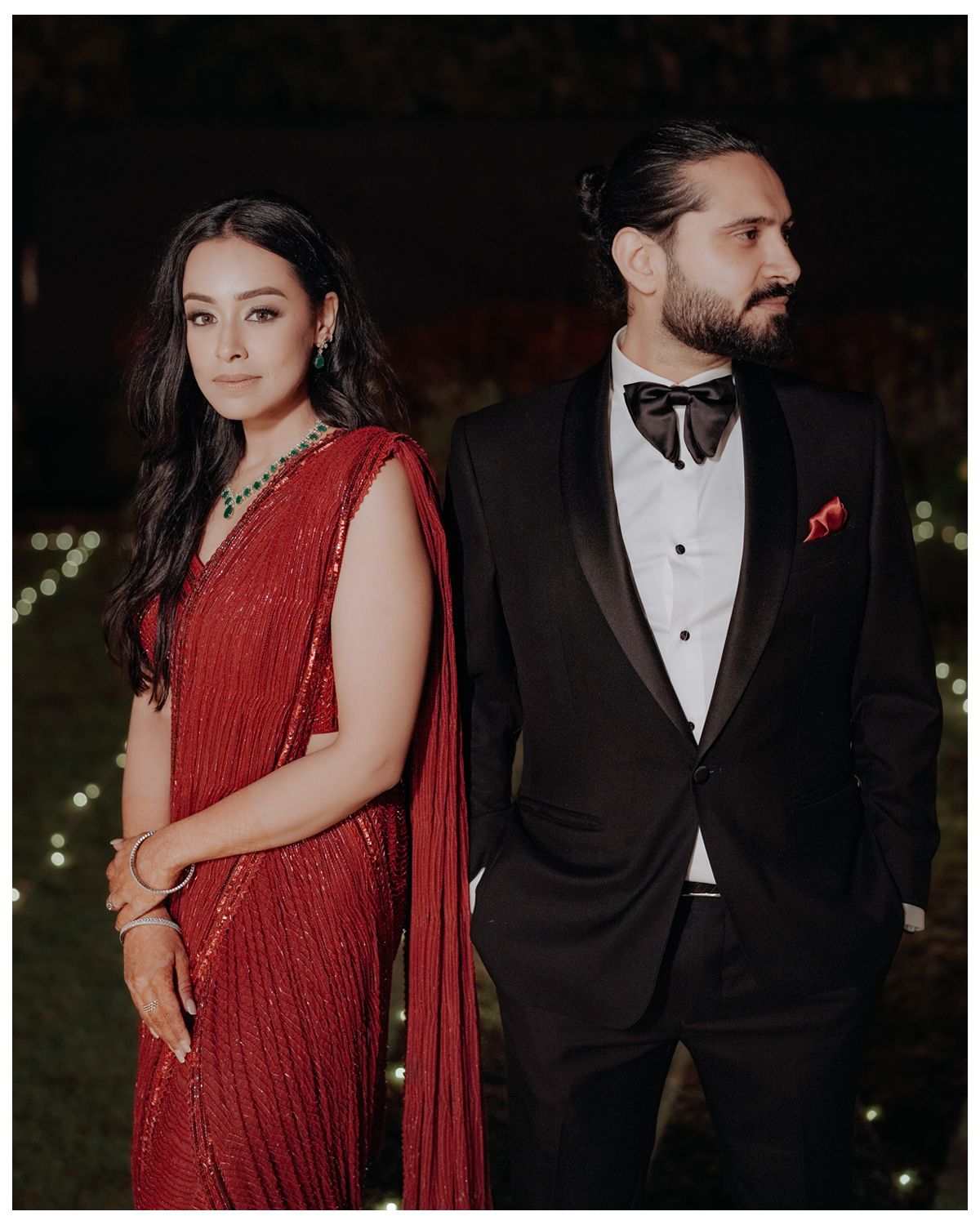 Couple in formal attire, with individual wearing red evening sari and person in black tuxedo standing side by side.