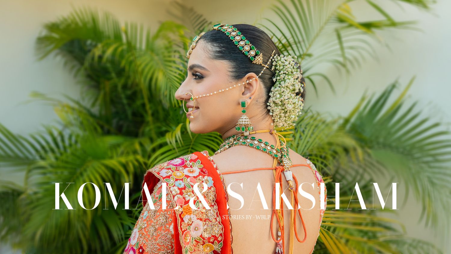 Indian bride in orange traditional attire with floral jewelry against tropical foliage.