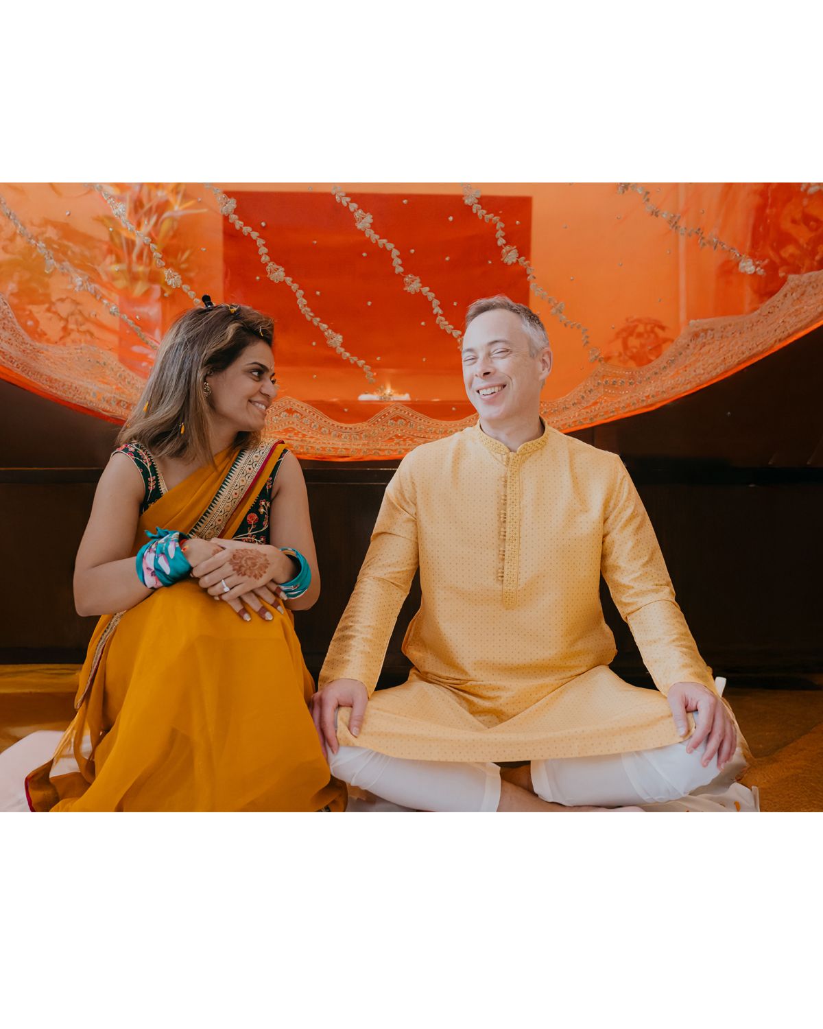 Couple in traditional Indian dress sitting on floor during cultural ceremony with orange backdrop.