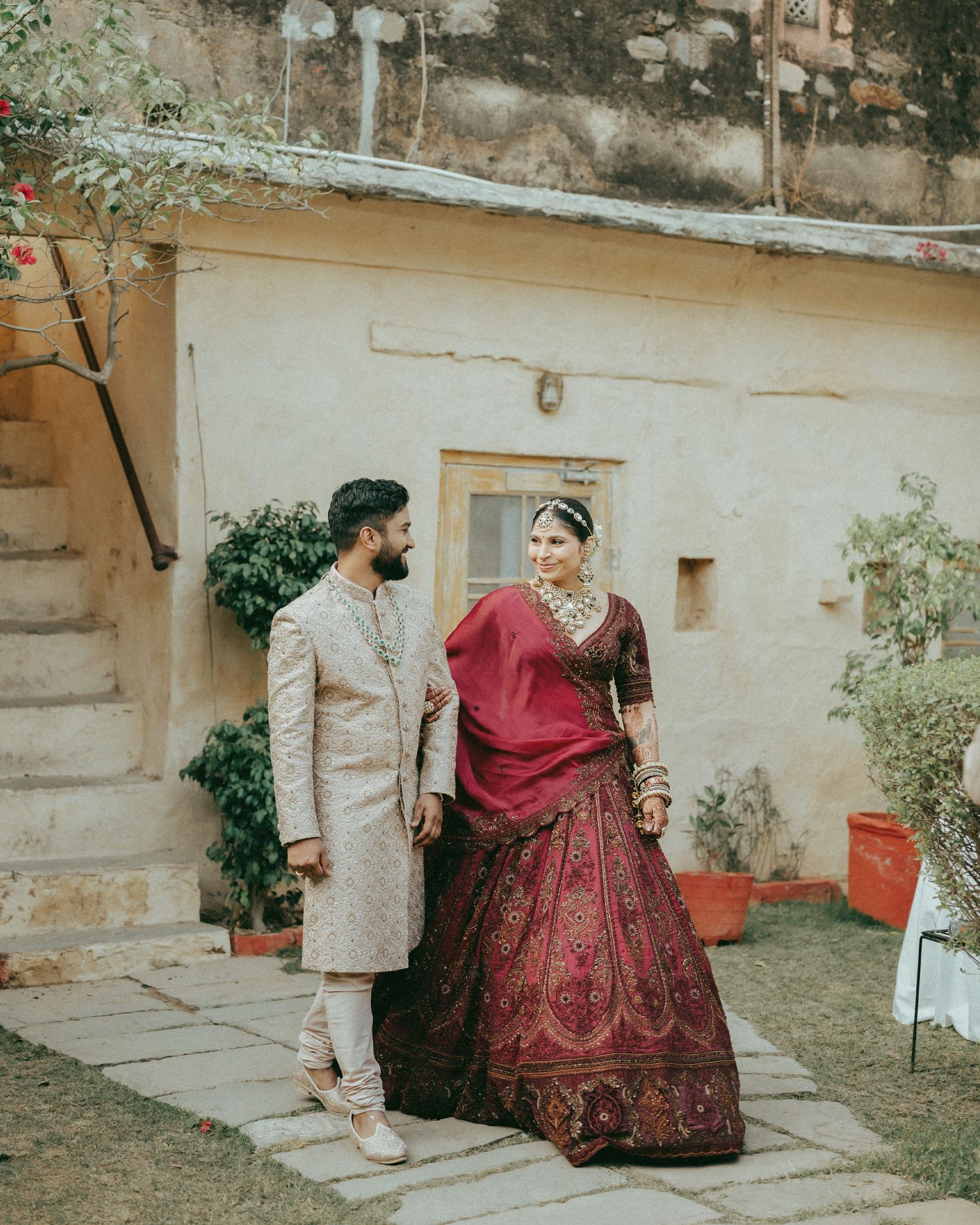 Indian bride in maroon lehenga and groom in beige sherwani share a romantic moment at a traditional wedding venue.