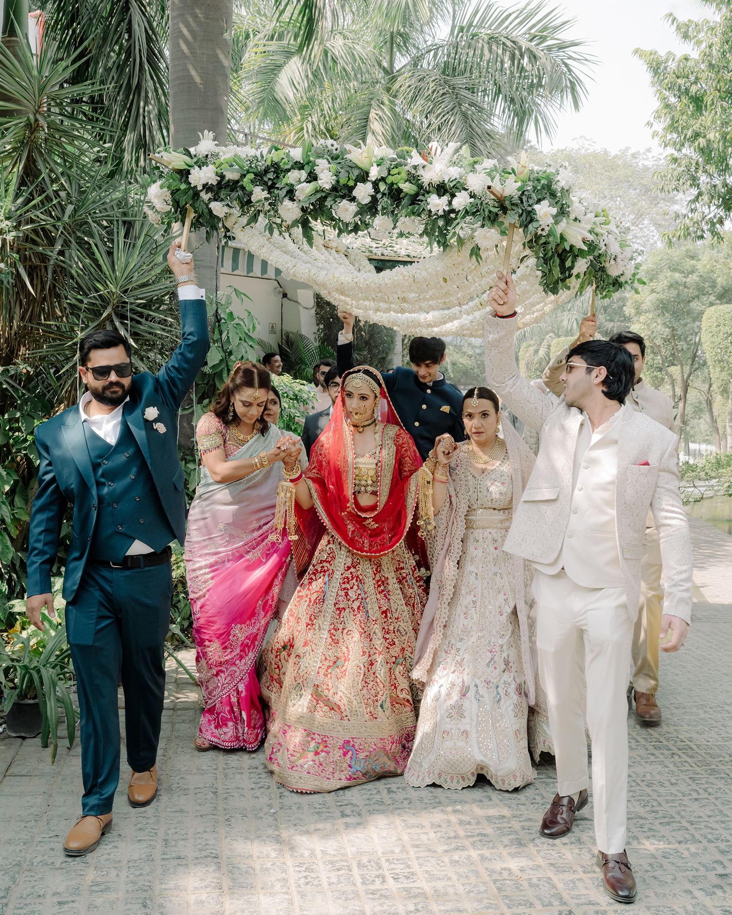 Bride in red lehenga walks under floral canopy held by groomsmen during traditional Indian wedding ceremony.