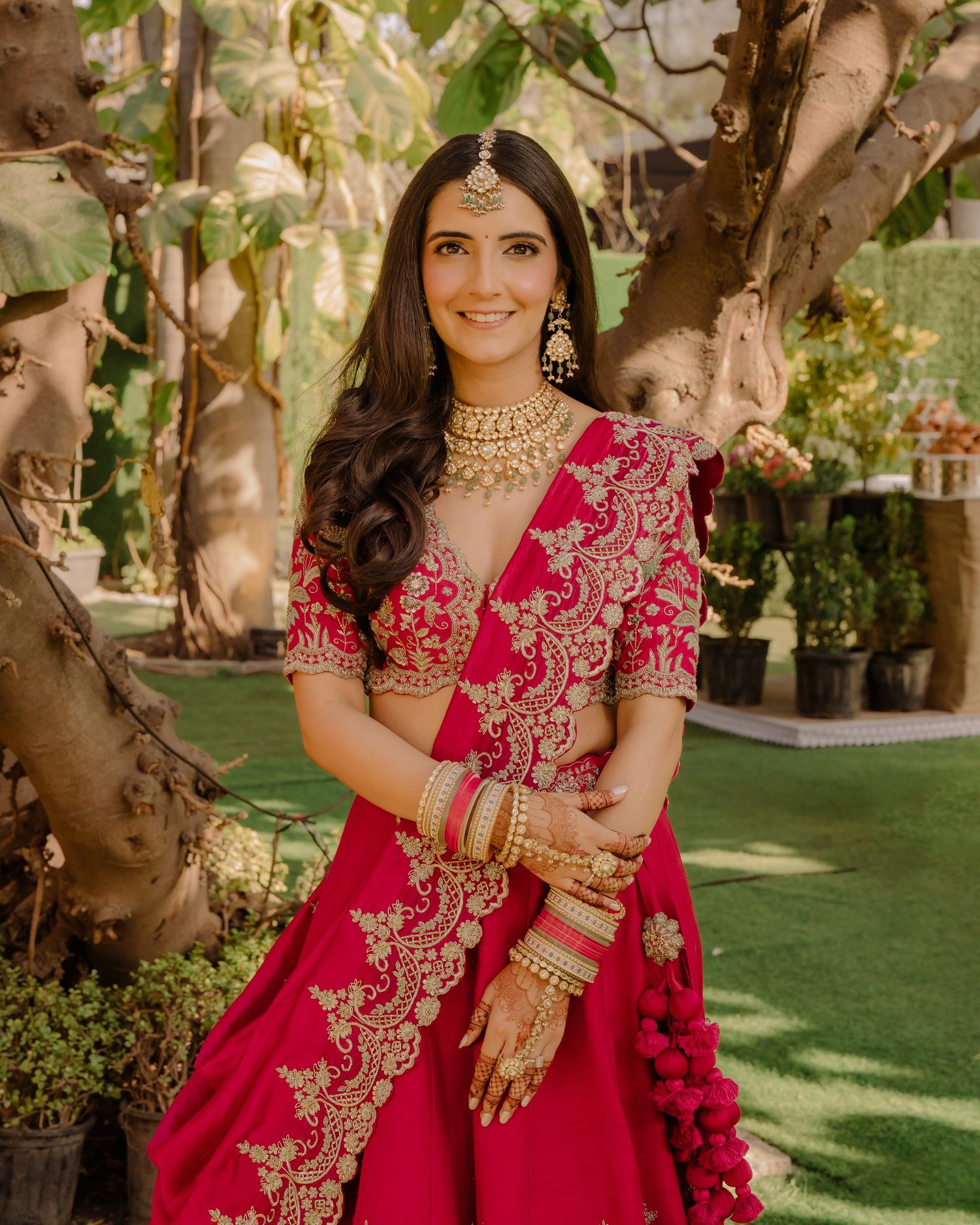 A bride in a red and gold embroidered lehenga with traditional jewelry smiles outdoors.
